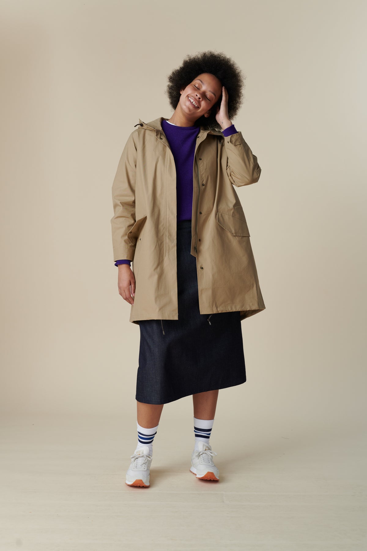 
            Front shot of a young, black female model with dark hair wearing long stone coloured parka. The parka is styled with lambswool crew neck in dark purple and midi skirt in indigo with white Walsh trainer.