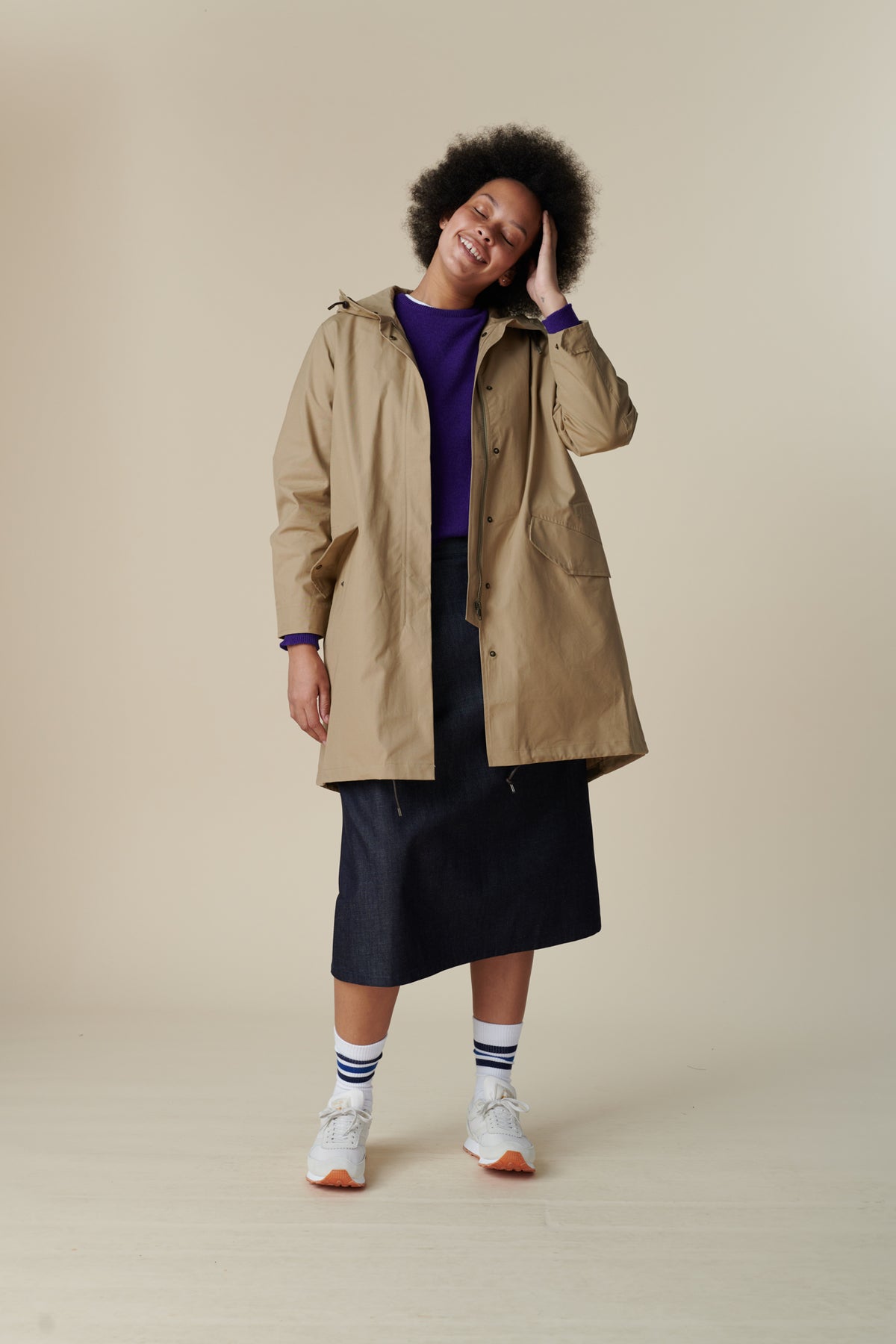 Front shot of a young, black female model with dark hair wearing long stone coloured parka. The parka is styled with lambswool crew neck in dark purple and midi skirt in indigo with white Walsh trainer.