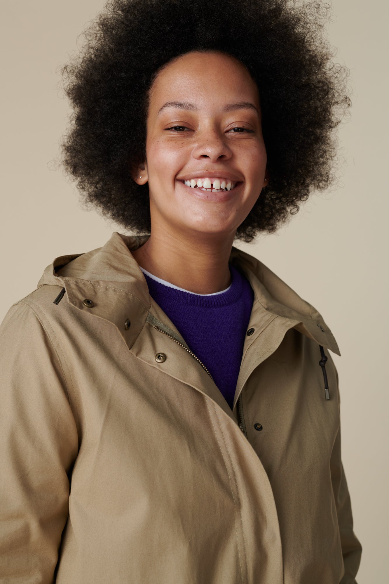 Shoulders up image of a young, black female model with dark hair wearing a long parka in stone over the women's lambswool crew neck in dark purple
