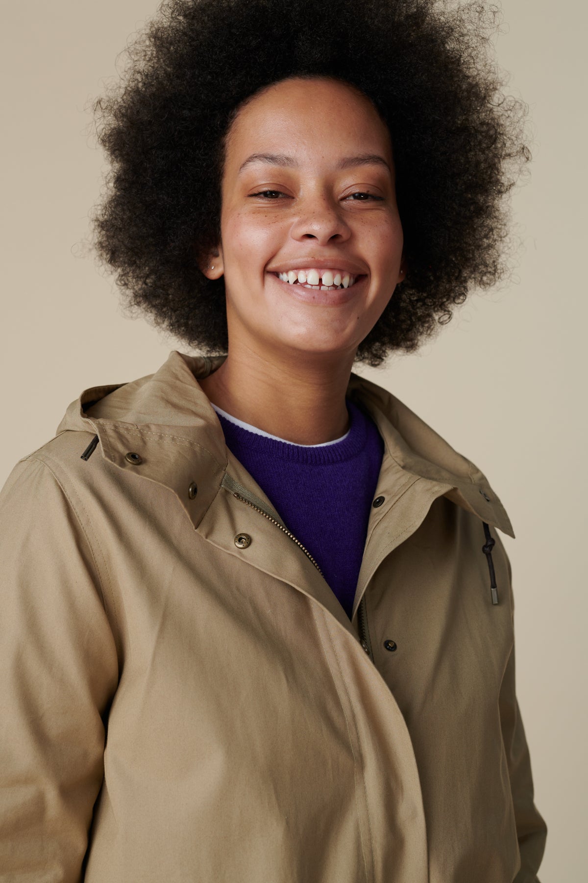Shoulders up image of a young, black female model with dark hair wearing a long parka in stone over the women's lambswool crew neck in dark purple