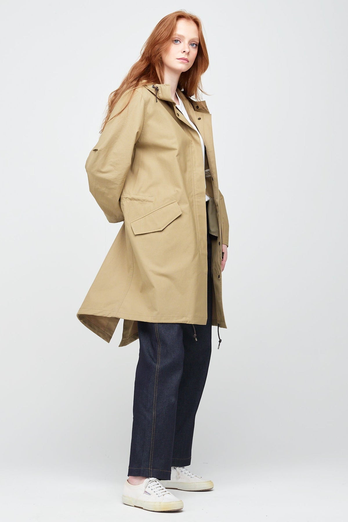 
            Side shot of a young, white female model with ginger hair wearing long stone coloured parka. The parka is styled with indigo jeans and a white trainer.