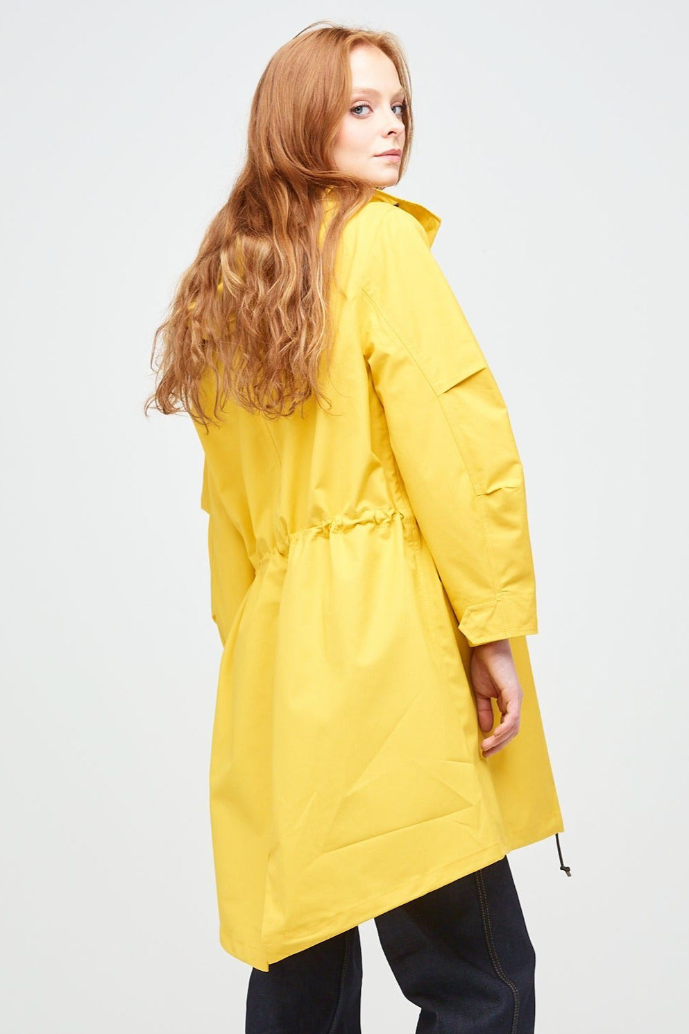 rear shot of a young, white female model with ginger hair wearing long yellow parka. The parka is styled with indigo jeans and a white trainer.