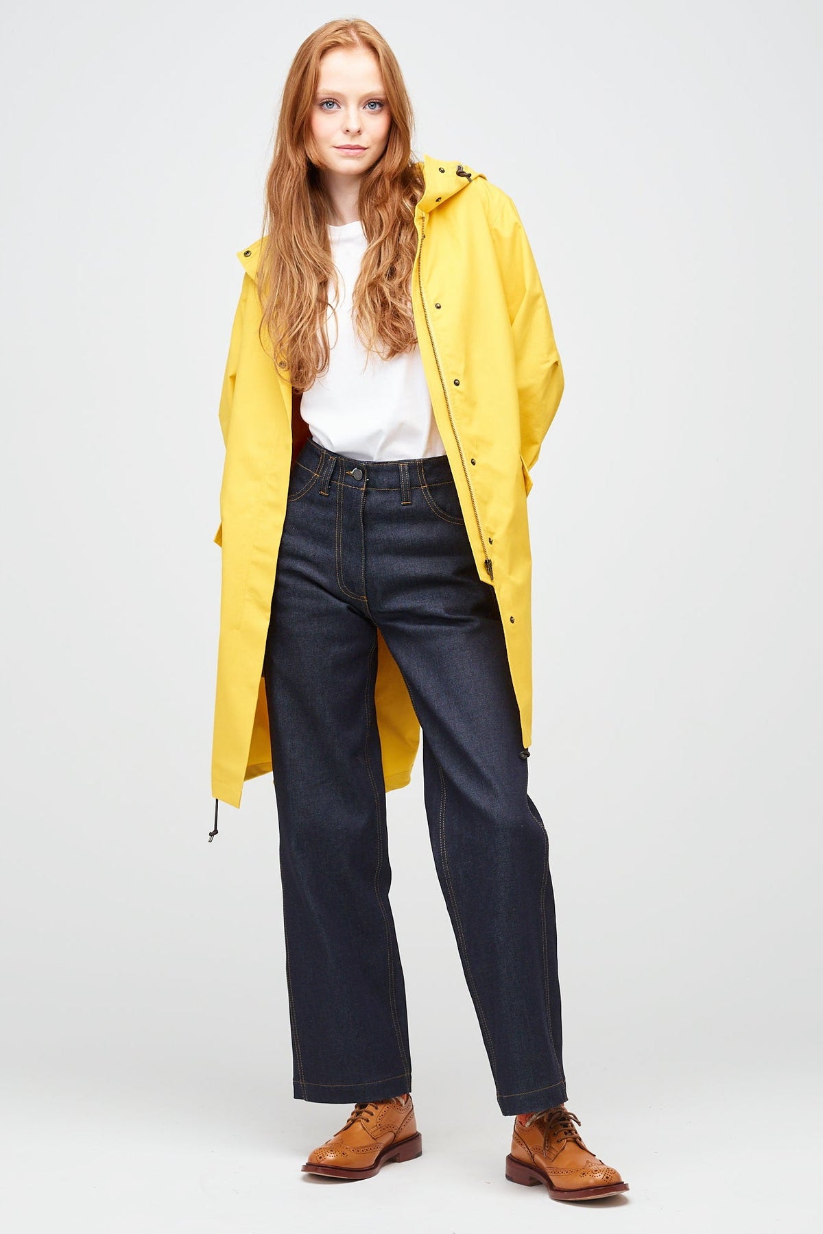 
            a young, white female model with ginger hair wearing long yellow parka unzipped. The parka is styled a white t-shirt, with indigo jeans and a white trainer.
