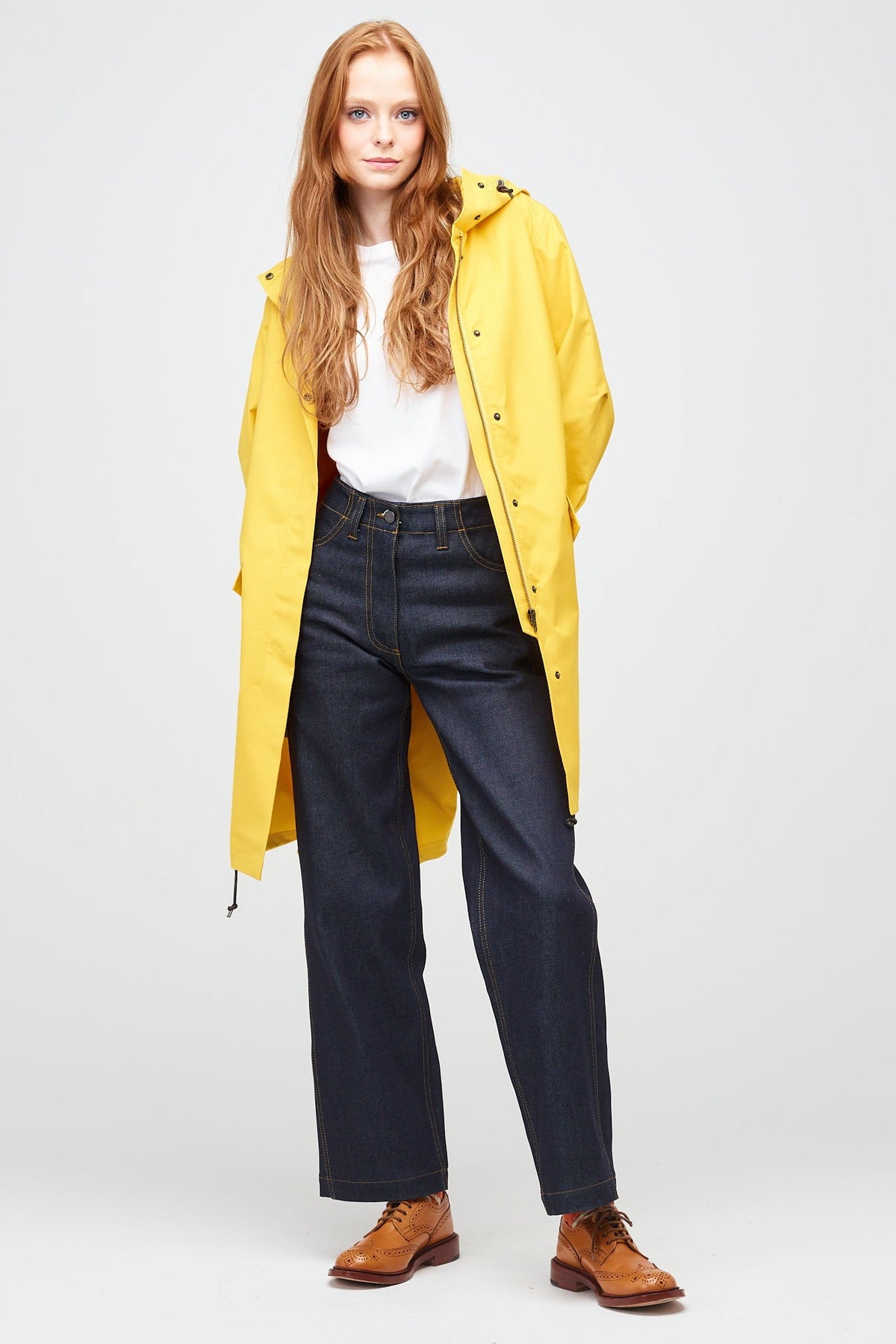 a young, white female model with ginger hair wearing long yellow parka unzipped. The parka is styled a white t-shirt, with indigo jeans and a white trainer.