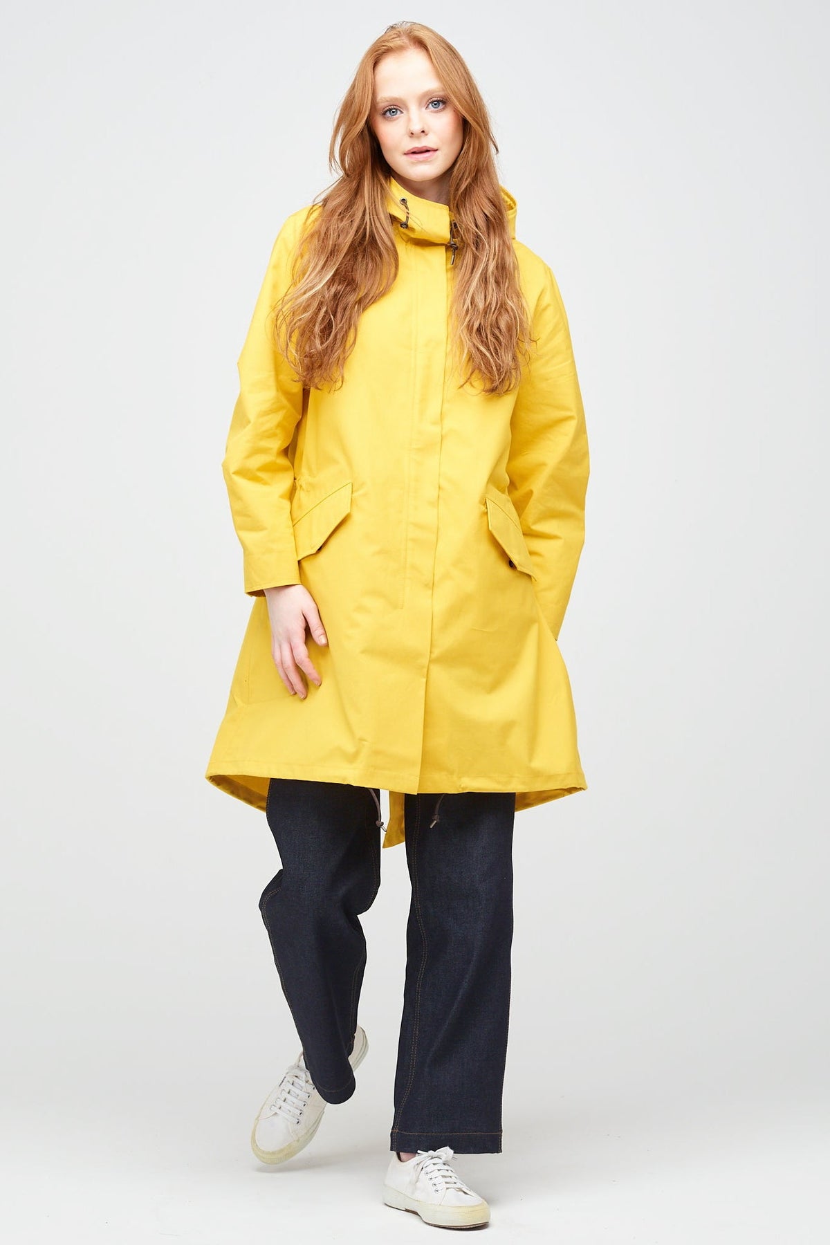 
            a young, white female model with ginger hair wearing long yellow parka fully zipped up. The parka is styled with indigo jeans and a white trainer.