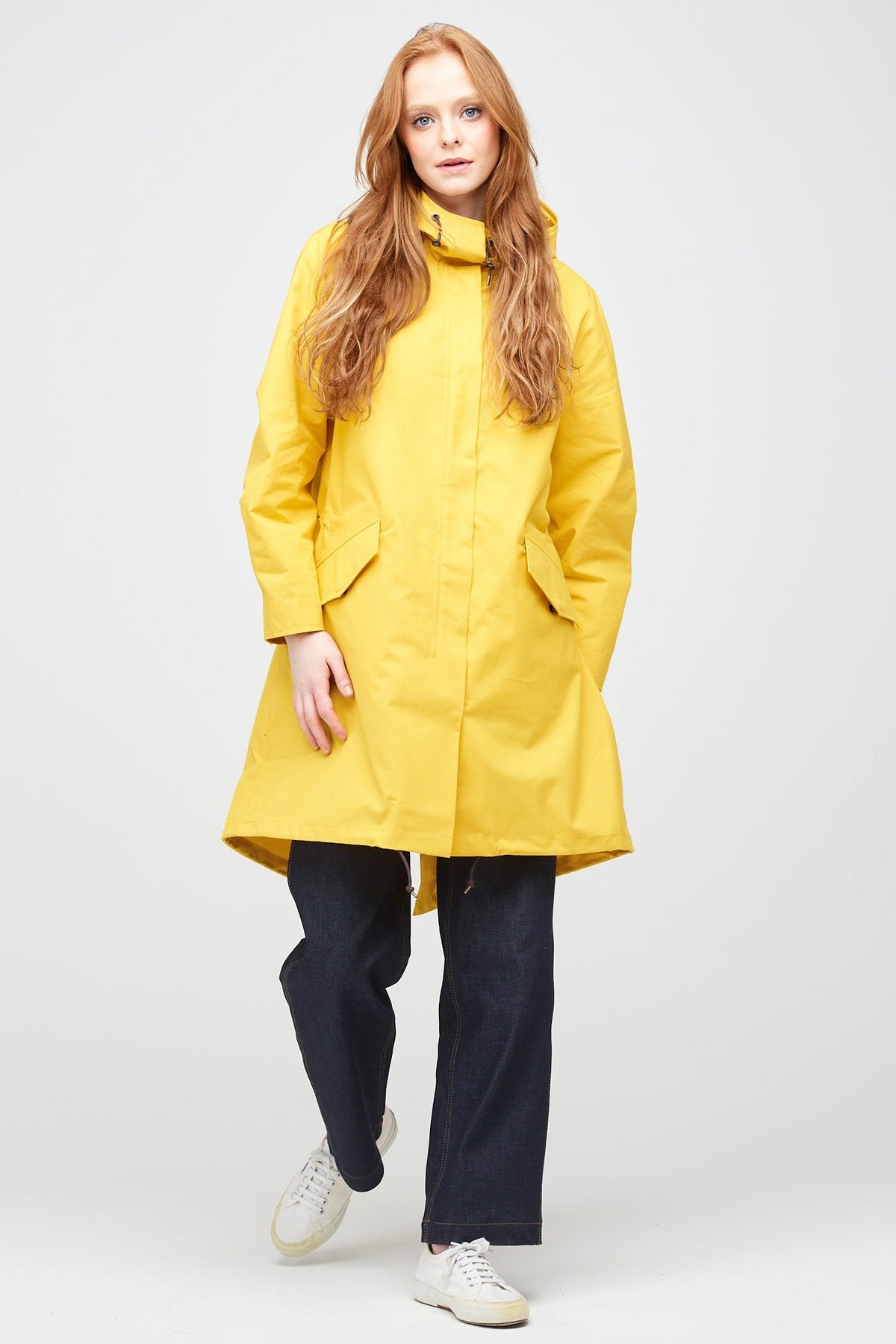 a young, white female model with ginger hair wearing long yellow parka fully zipped up. The parka is styled with indigo jeans and a white trainer.