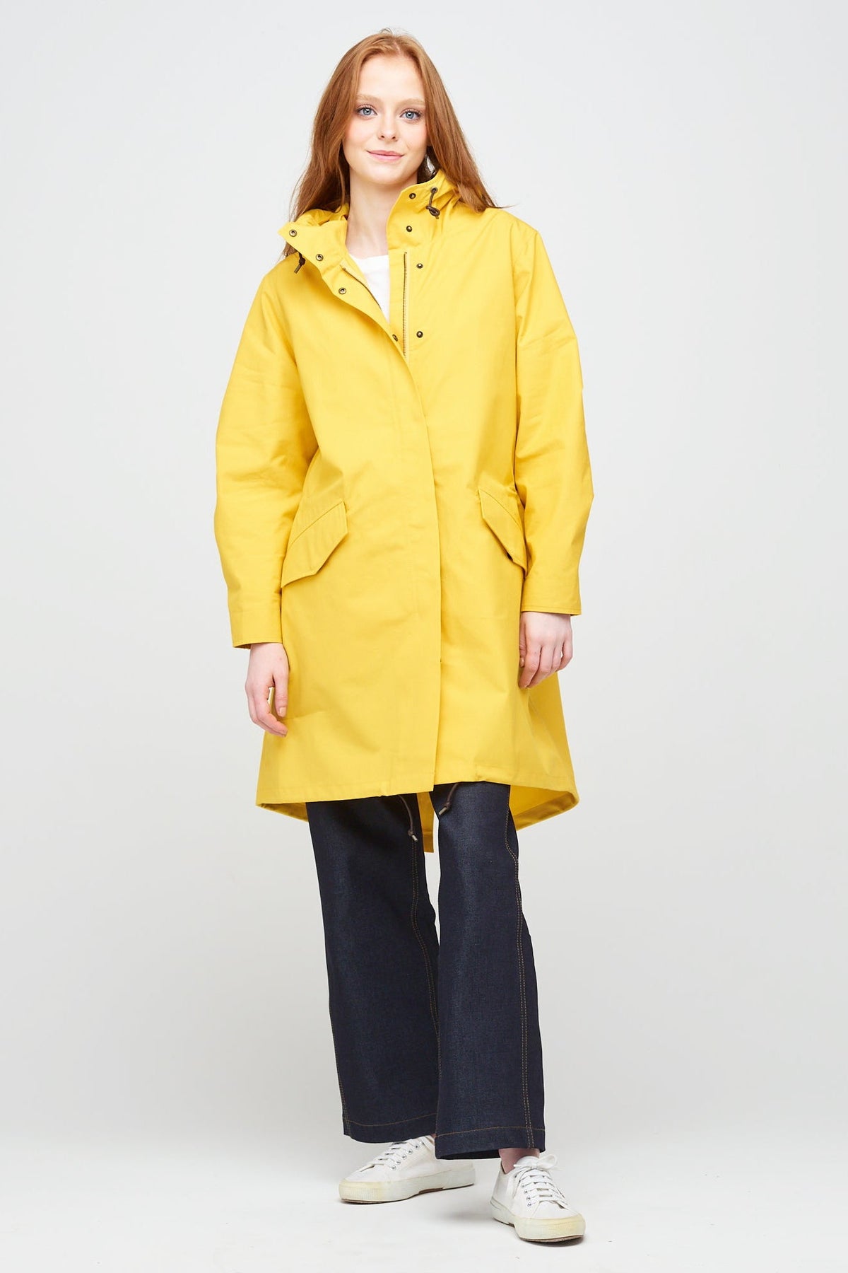 a young, white female model with ginger hair wearing long yellow parka partially zipped up. The parka is styled with indigo jeans and a white trainer.