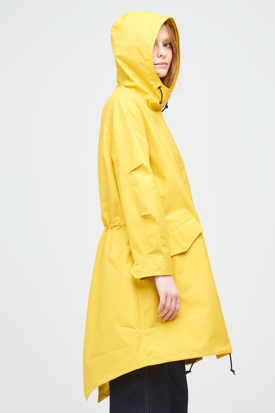 Side shot of a young, white female model with ginger hair wearing long yellow parka with the hood up. The parka is styled with indigo jeans and a white trainer.