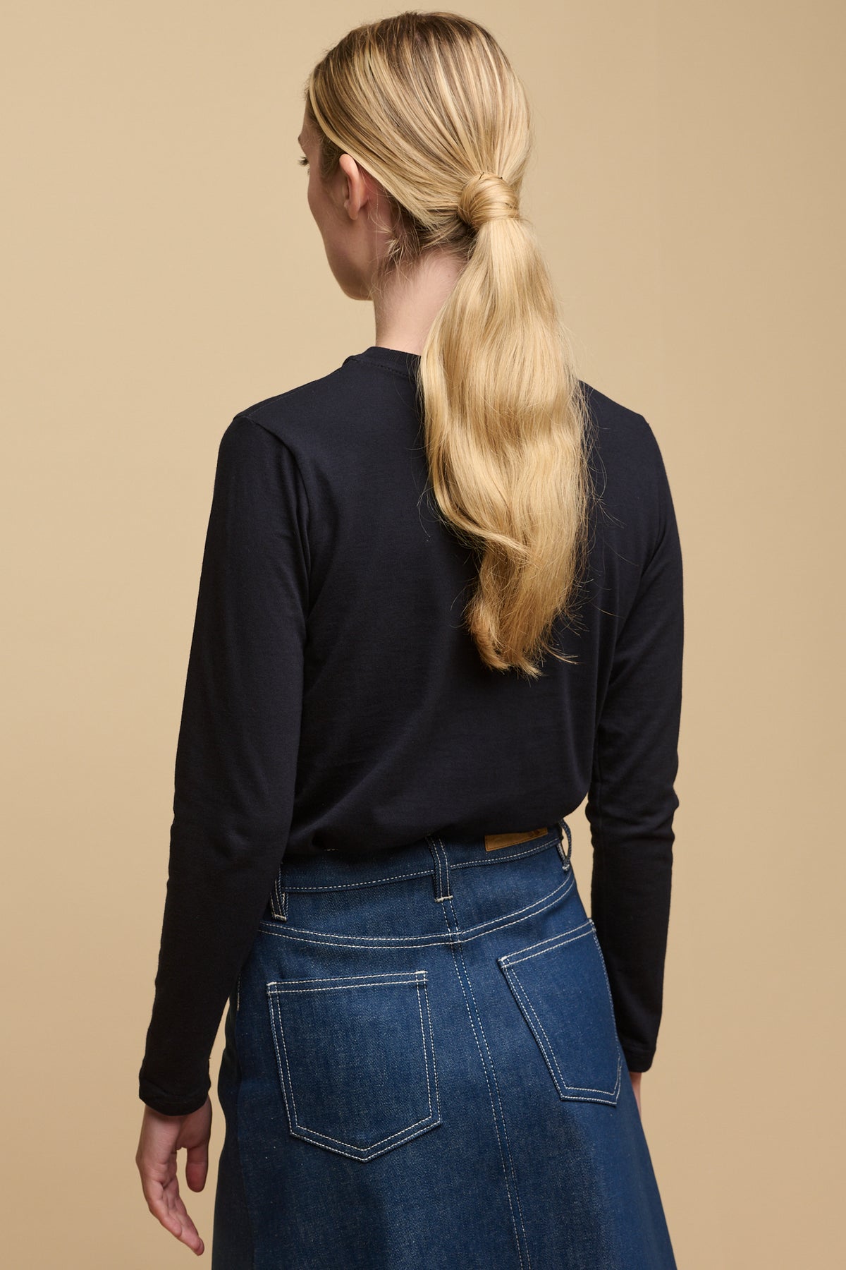 
            The back of female with long blonde hair tied in low ponytail wearing long sleeve t-shirt in navy tucked into Bobbie denim midi skirt in blue