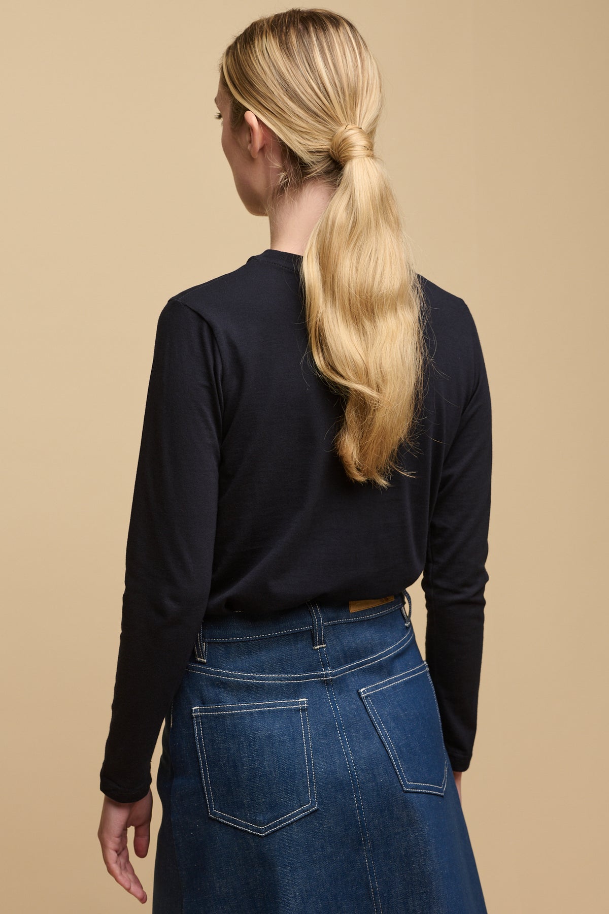 The back of female with long blonde hair tied in low ponytail wearing long sleeve t-shirt in navy tucked into Bobbie denim midi skirt in blue