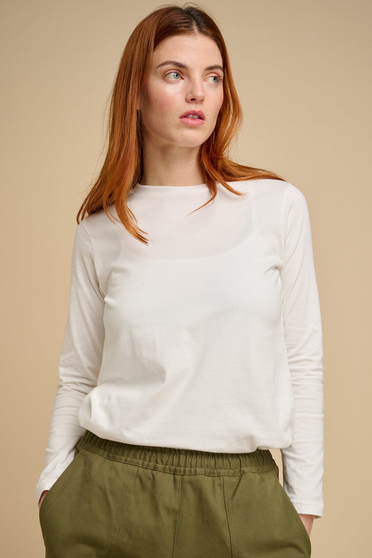 Woman wearing a bone long-sleeve t shirt and green pants against a beige background.