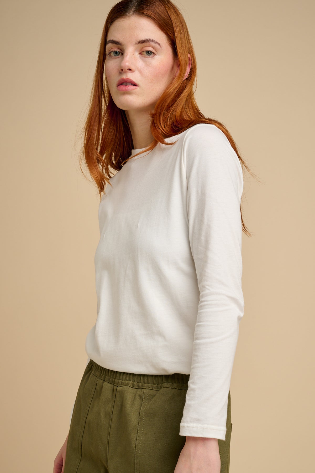 Woman wearing a bone long-sleeve t shirt and green pants against a beige background.