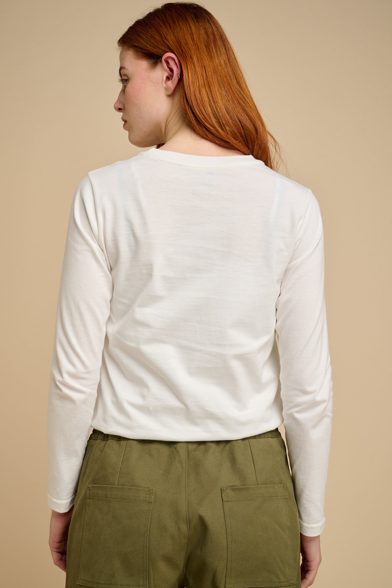 Woman wearing a bone long-sleeve t shirt and green pants against a beige background.