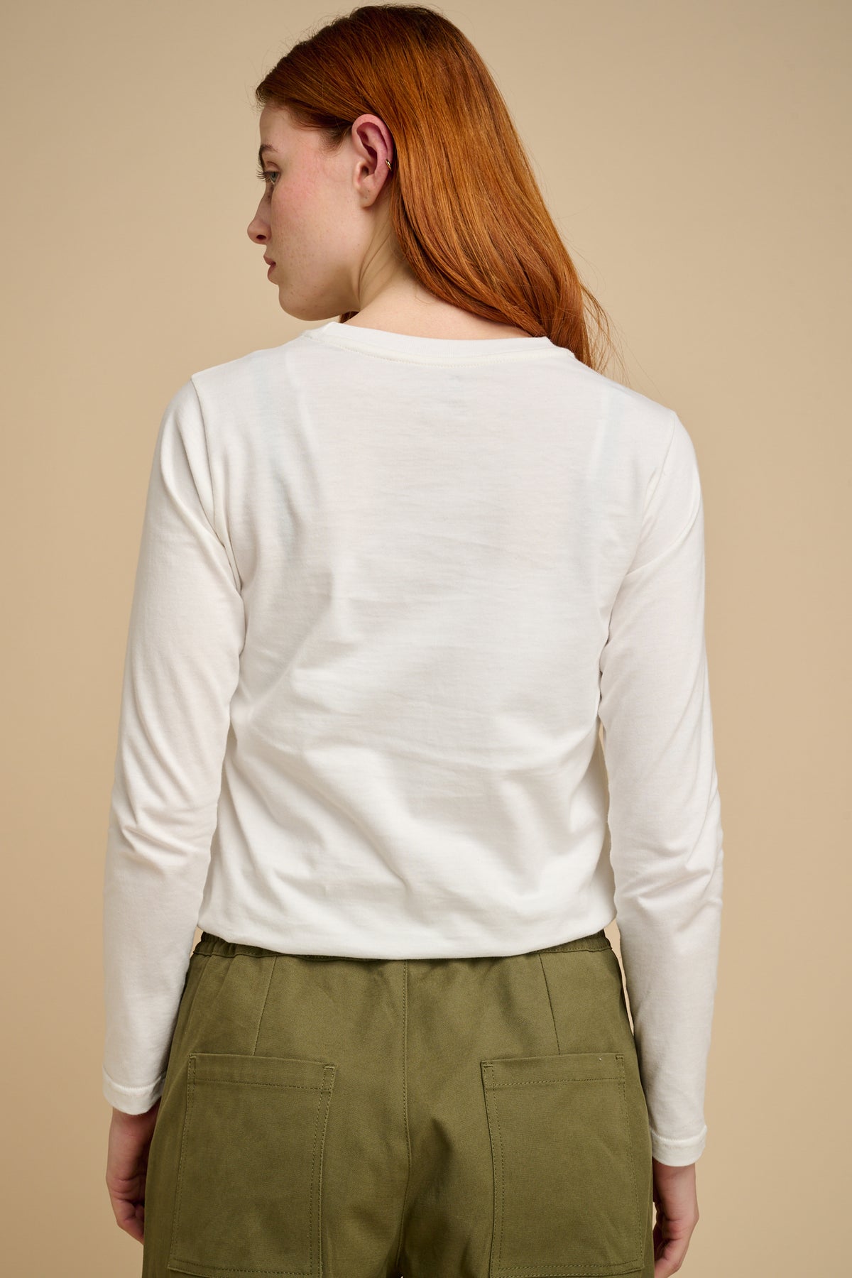 Woman wearing a bone long-sleeve t shirt and green pants against a beige background.