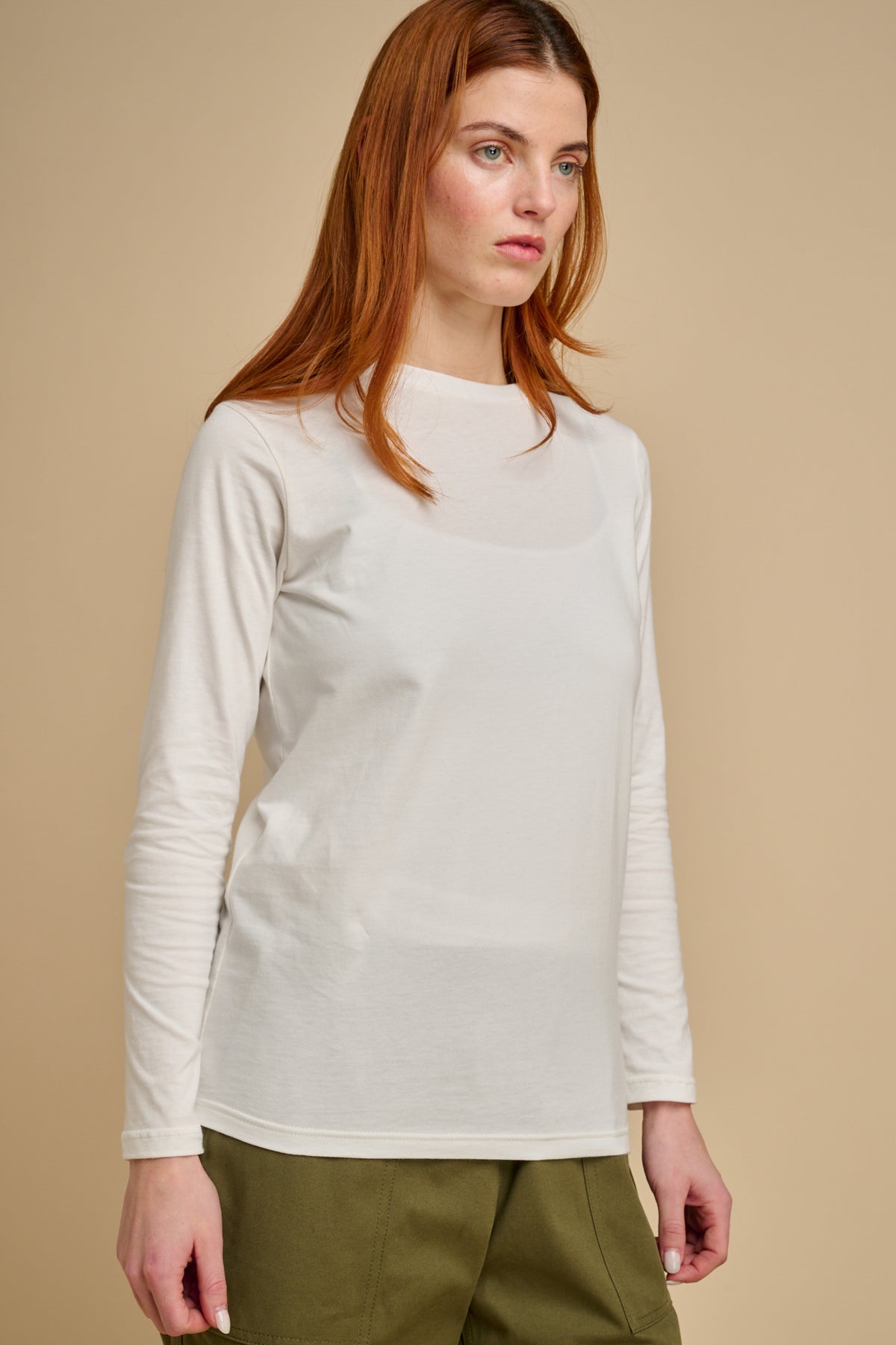 Woman wearing a bone long-sleeve t shirt and green pants against a beige background.
