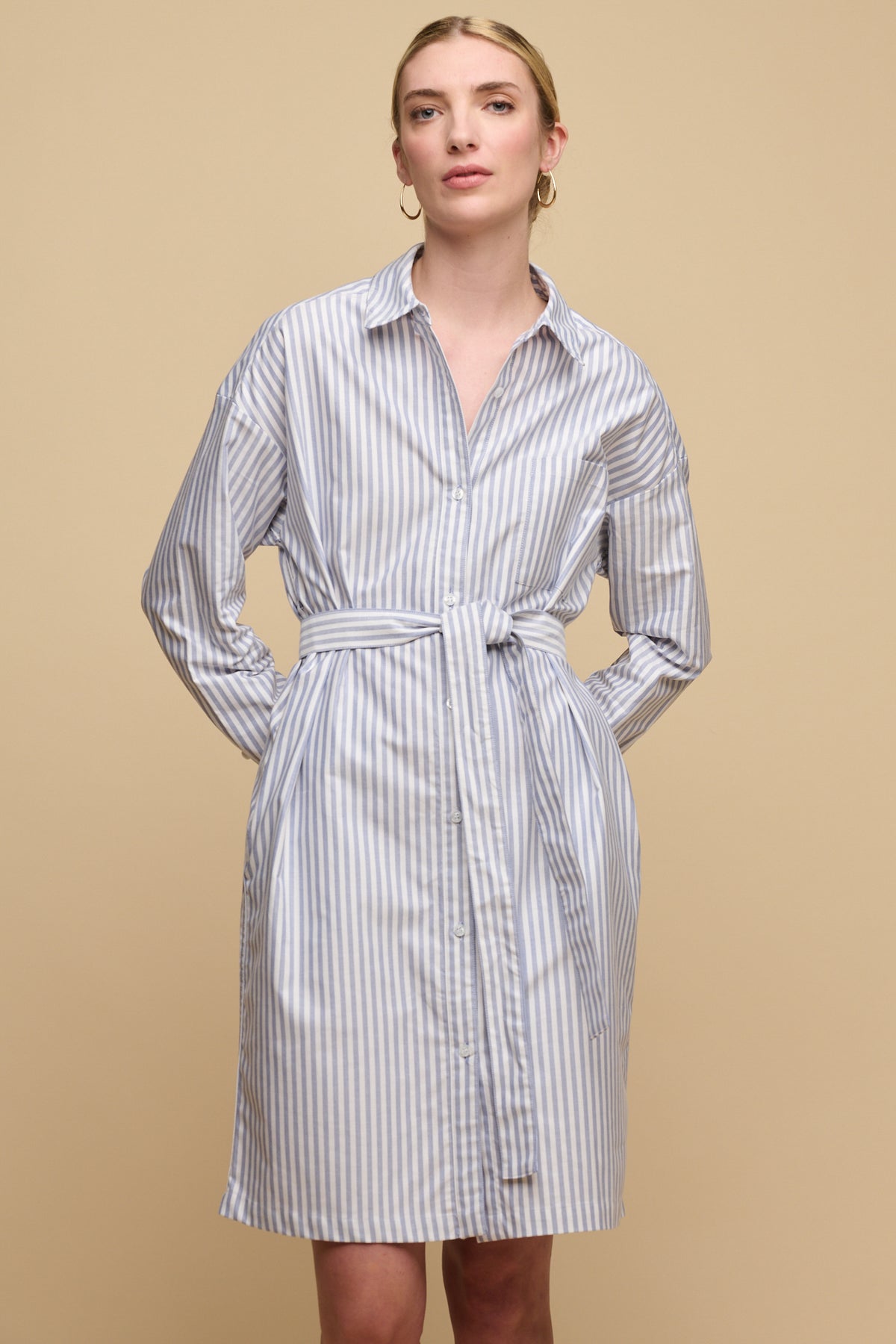 Thigh up image of white female with blonde hair tied back wearing belted midi cotton shirt dress in white blue stripes