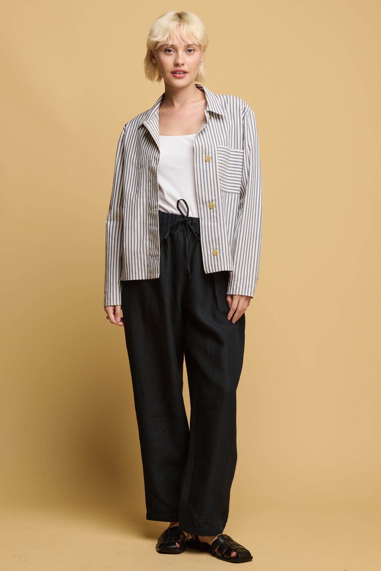 front of blonde female wearing community clothing lorna  elasticated waist linen rousers in navy paired with white camisole tucked in with quarry bank blue stripe overshirt with black sandals.