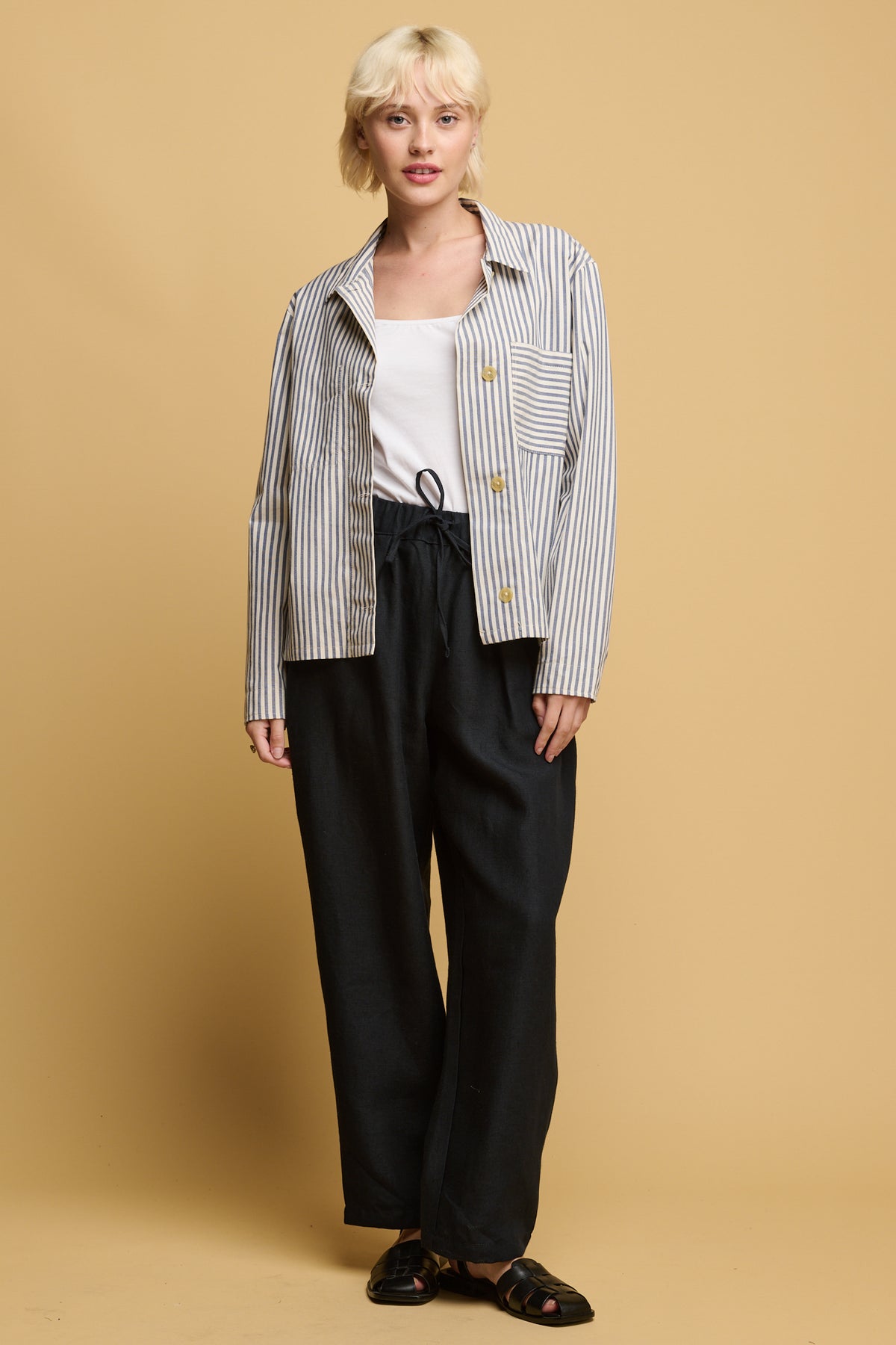 front of blonde female wearing community clothing lorna elasticated waist linen rousers in navy paired with white camisole tucked in with quarry bank blue stripe overshirt with black sandals.