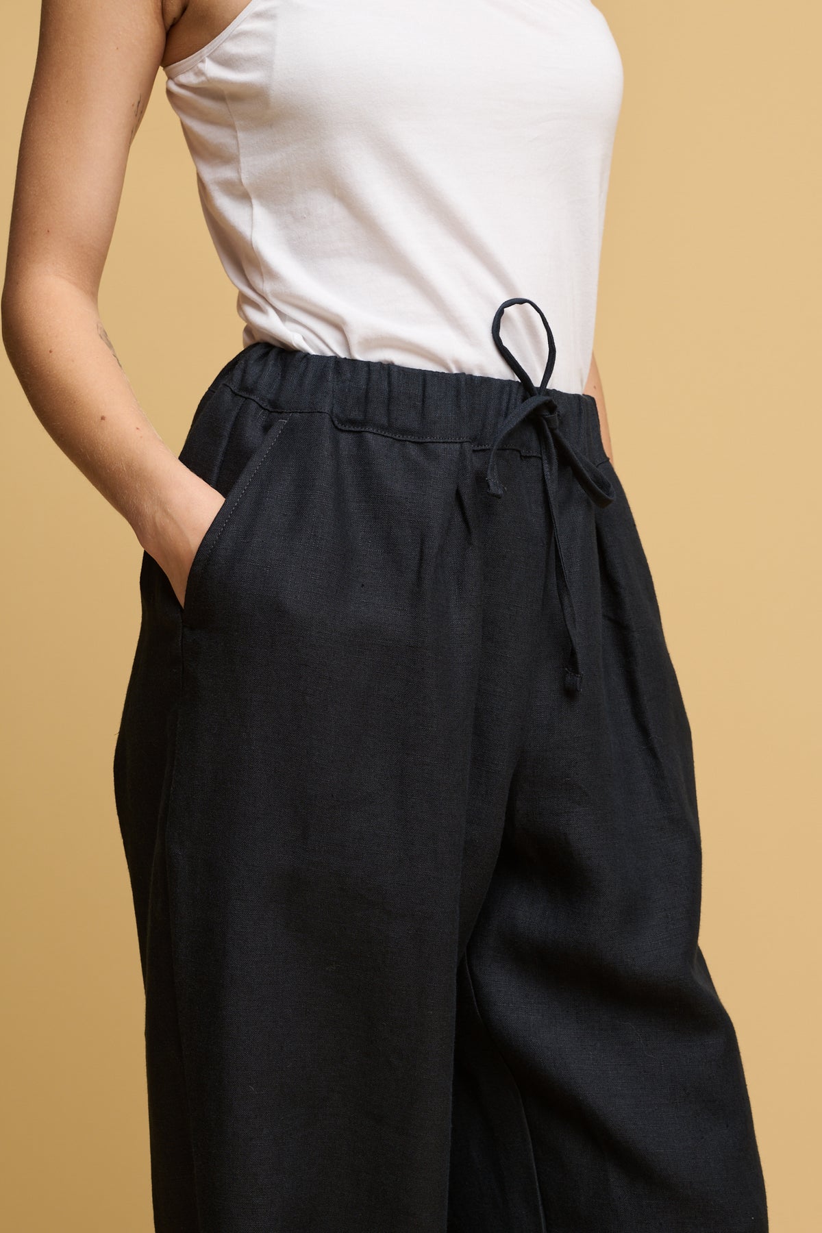 Close up of the shoulder to thigh of female with her hands in the pockets of lorna linen trousers with elasticated waist and adjustable drawstring tied into a bow.