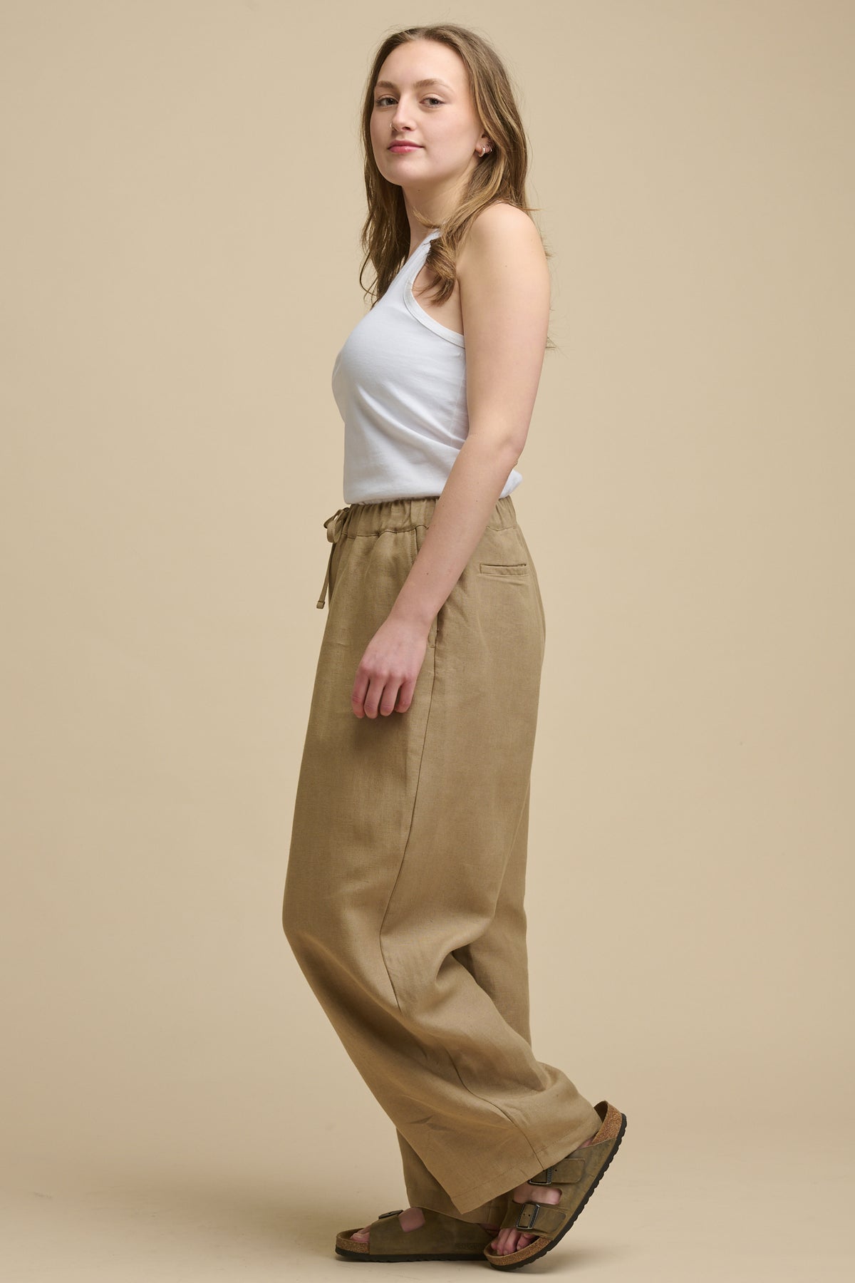 Full body image of the side of model wearing lorna elasticated waist linen trousers in truffle with drawstring waist tie and two back jet pockets wonr with racer back vest in white