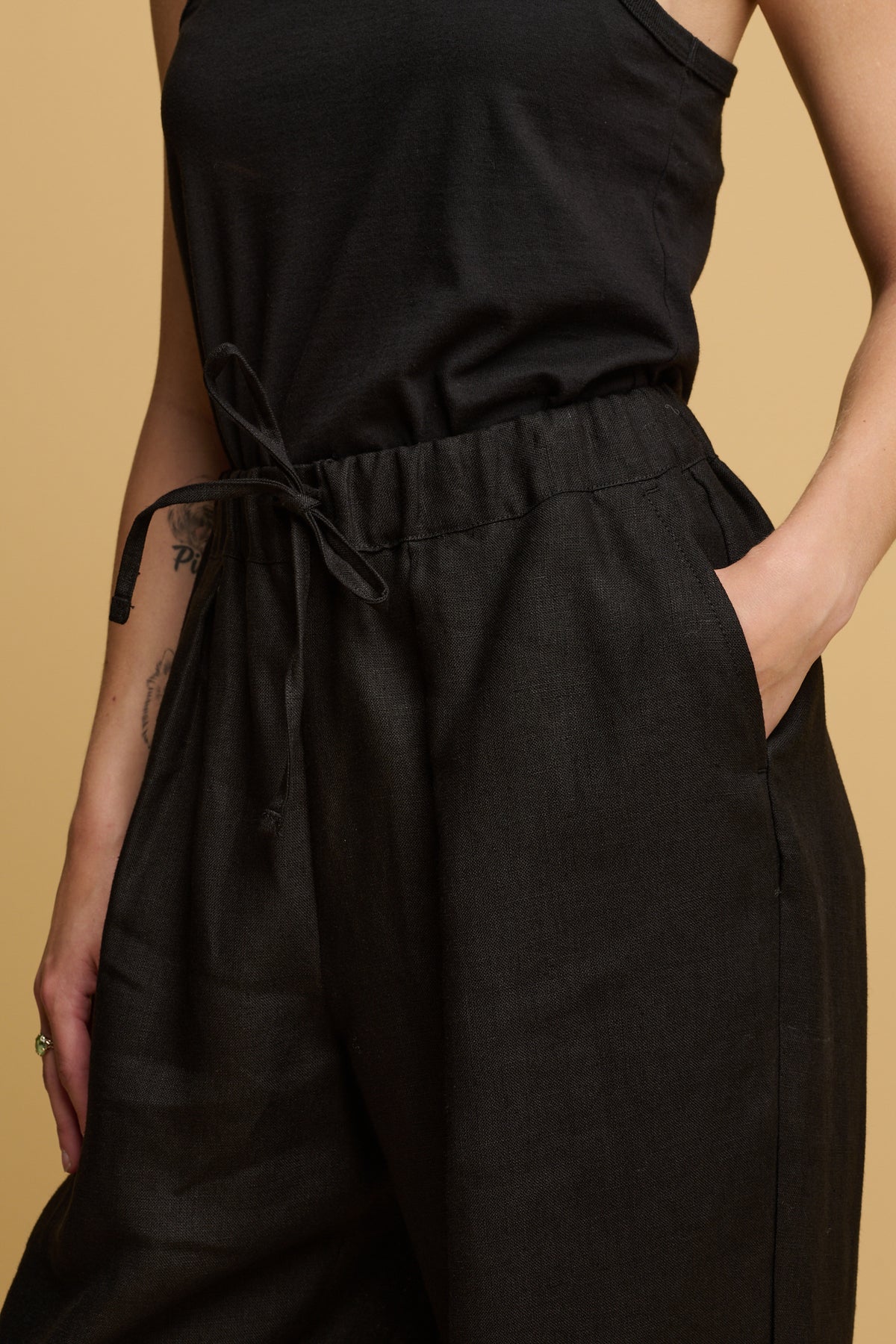 Close up of the front of female wearing lorna linen trousers in black with her hand in the front pocket and the drawstring through the waist tied in a bow