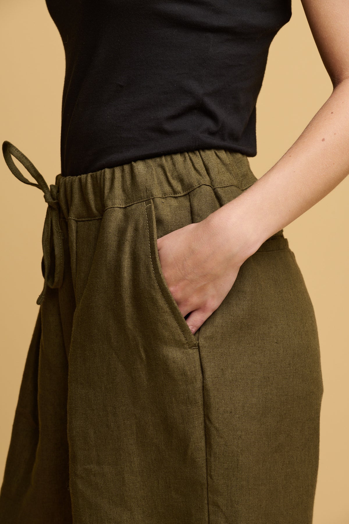 close up detail of white female wearing olive linen trousers with her hand in one of the front pockets, drawstring tied in a bow at the front of waistband.