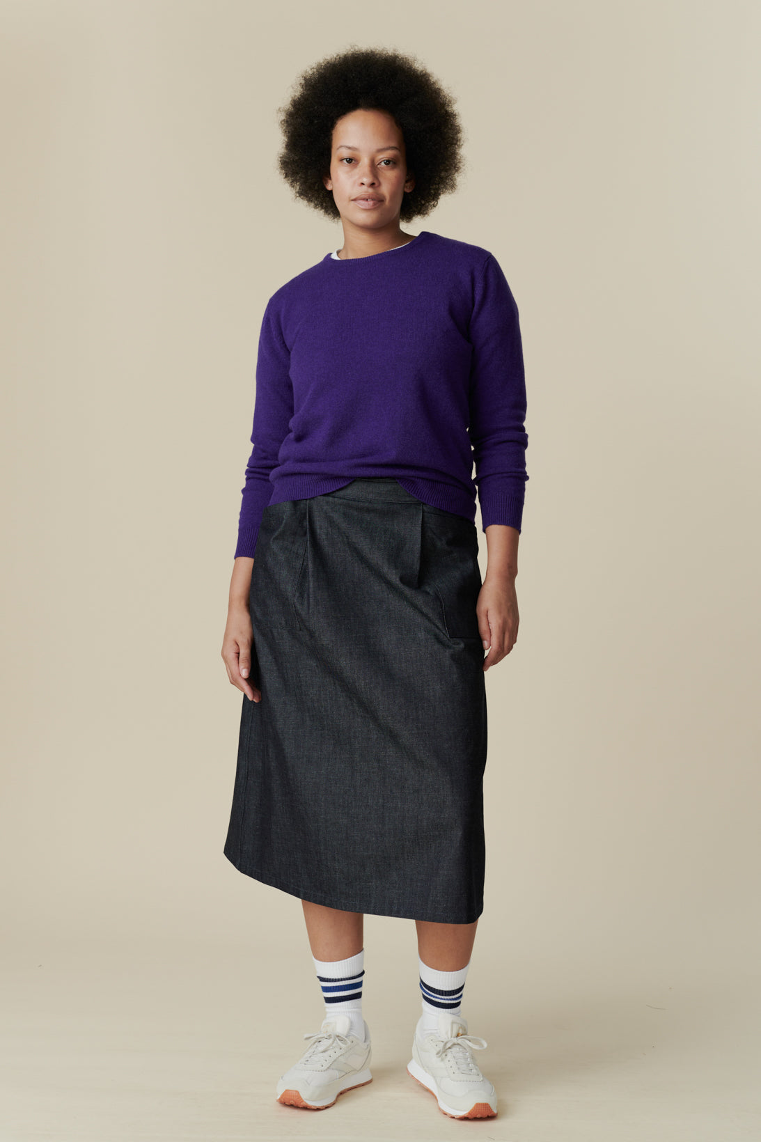 Full body image of the front of black female wearing midi skirt in denim paired with lambswool jumper and beacon trainers