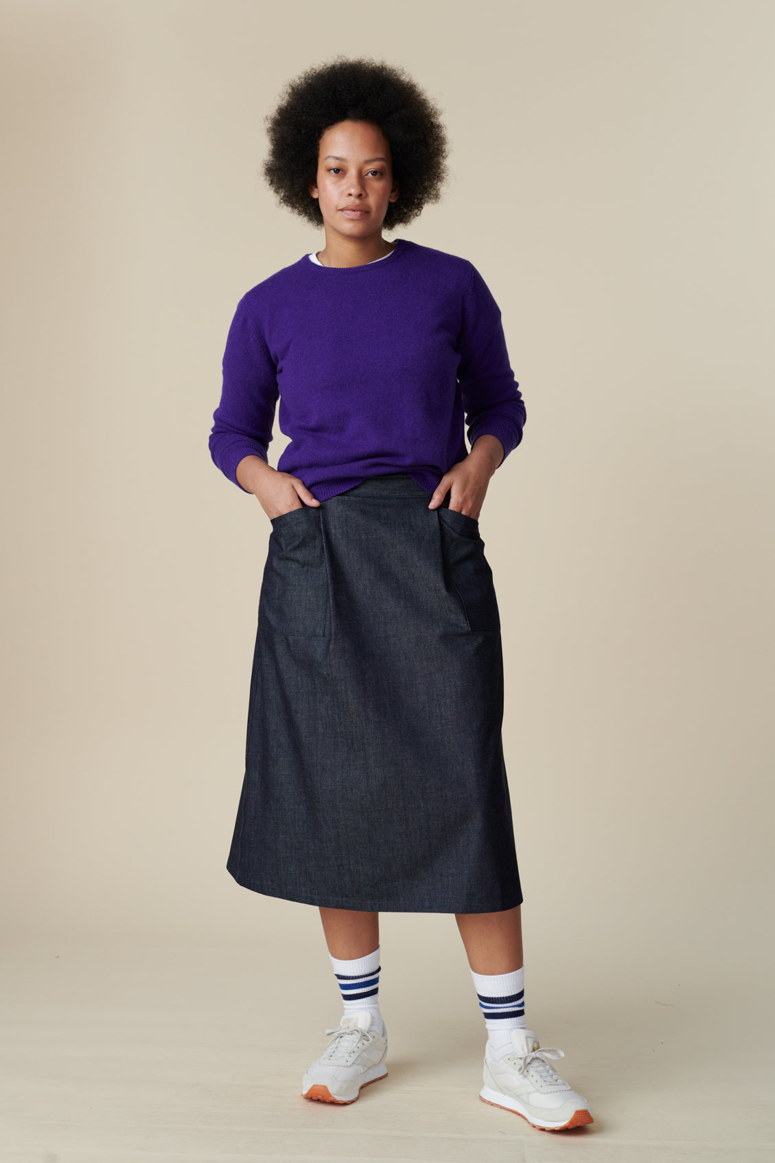 Full body image of the front of black model with afro wearing midi skirt in denim with her hands in the two front pockets of skirt worn with lambswool jumper in purple and sports socks with trainers
