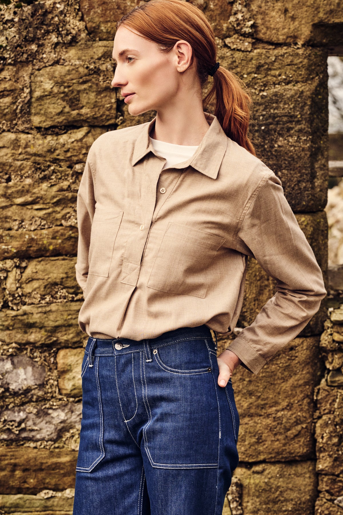 Woman wearing a beige shirt and blue jeans against a stone wall.