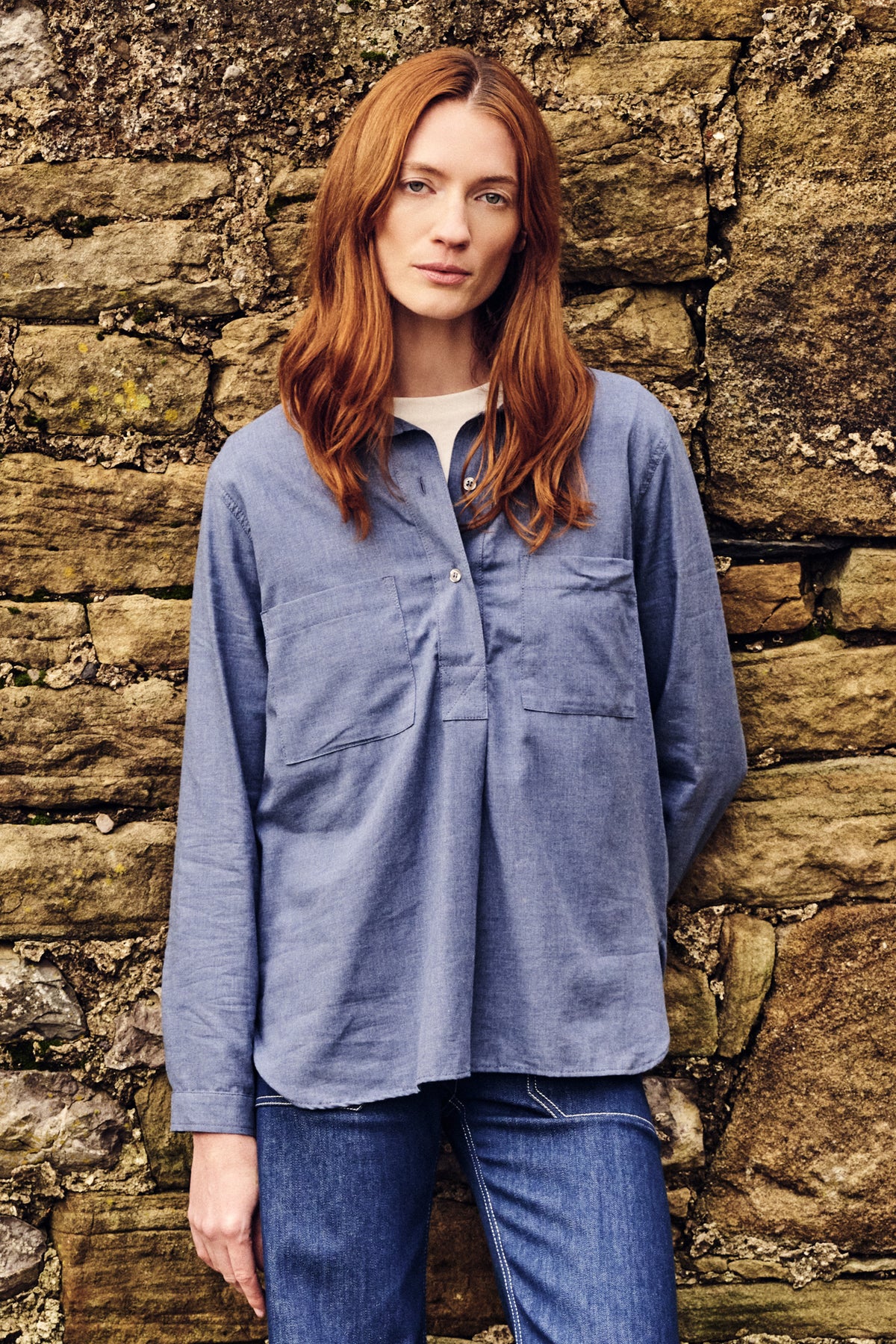 Woman wearing a blue shirt and jeans standing against a stone wall.