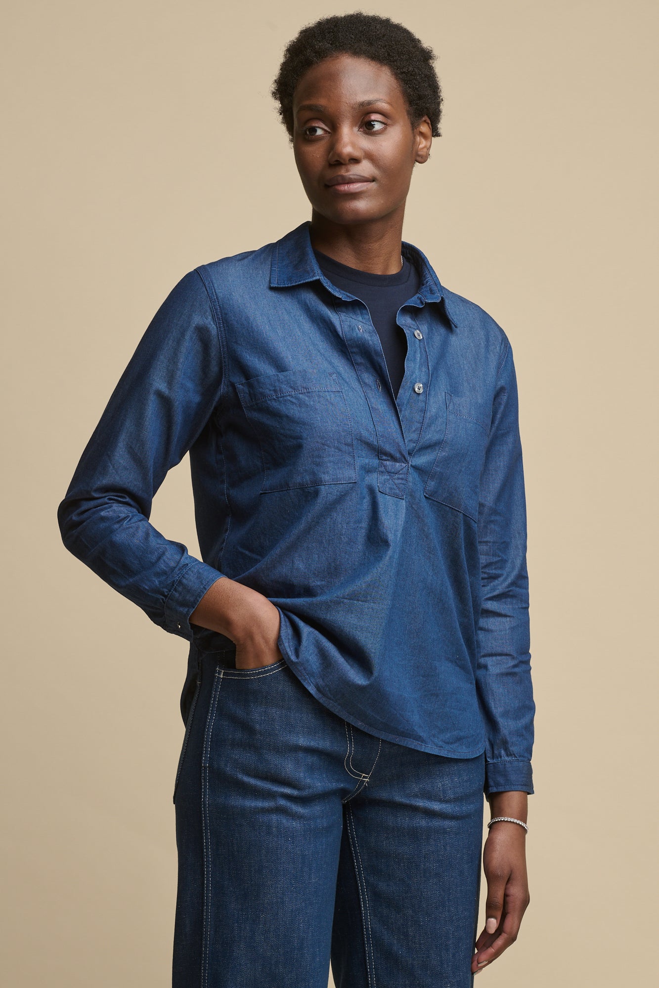 Thigh up image of female wearing Olivia half placket lightweight denim shirt in indigo, with button fastenings undone over crew neck t shirt in navy