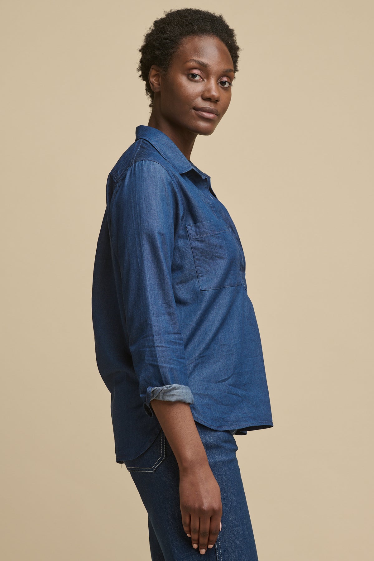 Thigh up image of the side of female wearing denim shirt in indigo with sleeves rolled up paired with blue jeans