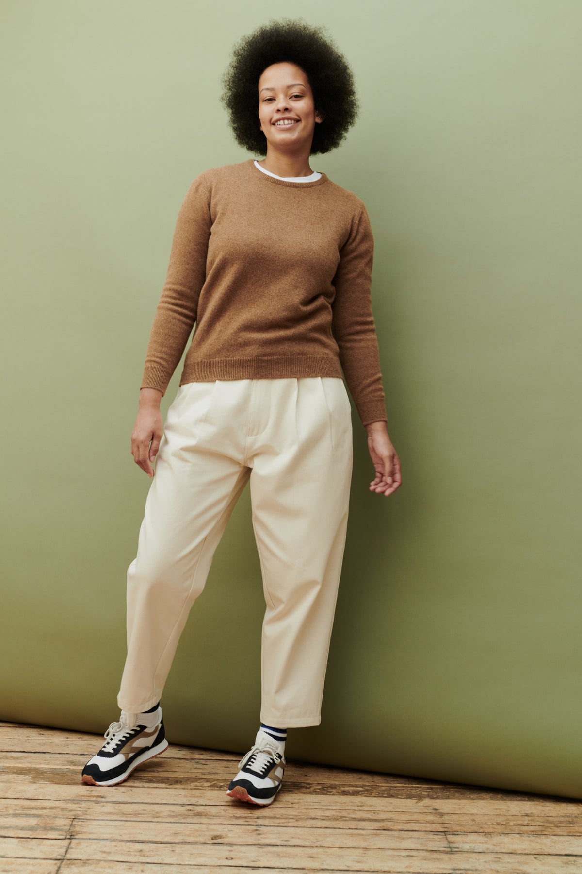 
            Full body shot of black female with afro wearing women&#39;s pleated jean in ecru paired with dark camel lambswool crew neck jumper and Walsh x community clothing trainers