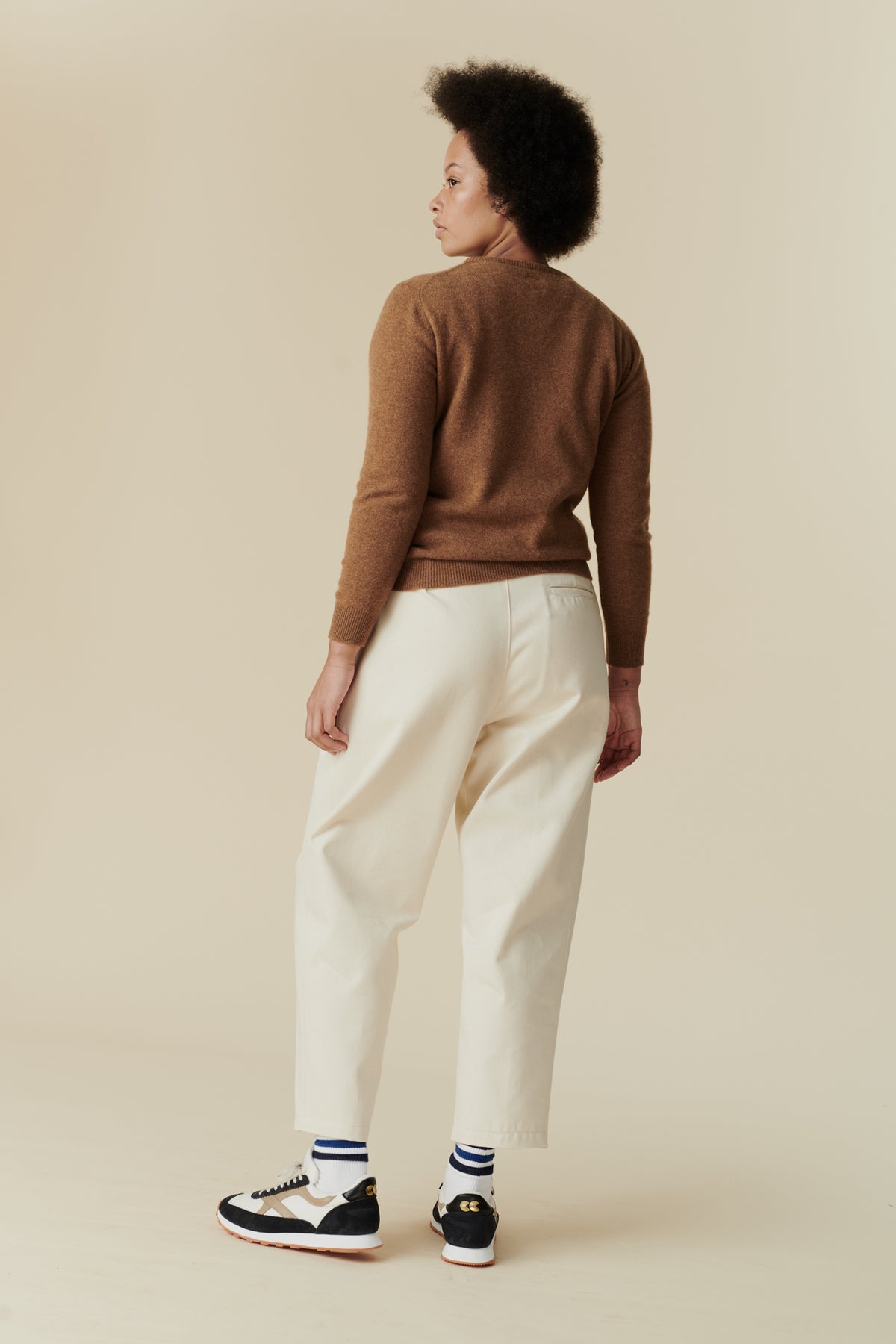 Full body shot of black female with afro wearing women's pleated jean in ecru paired with dark camel lambswool crew neck jumper and Walsh x community clothing trainers