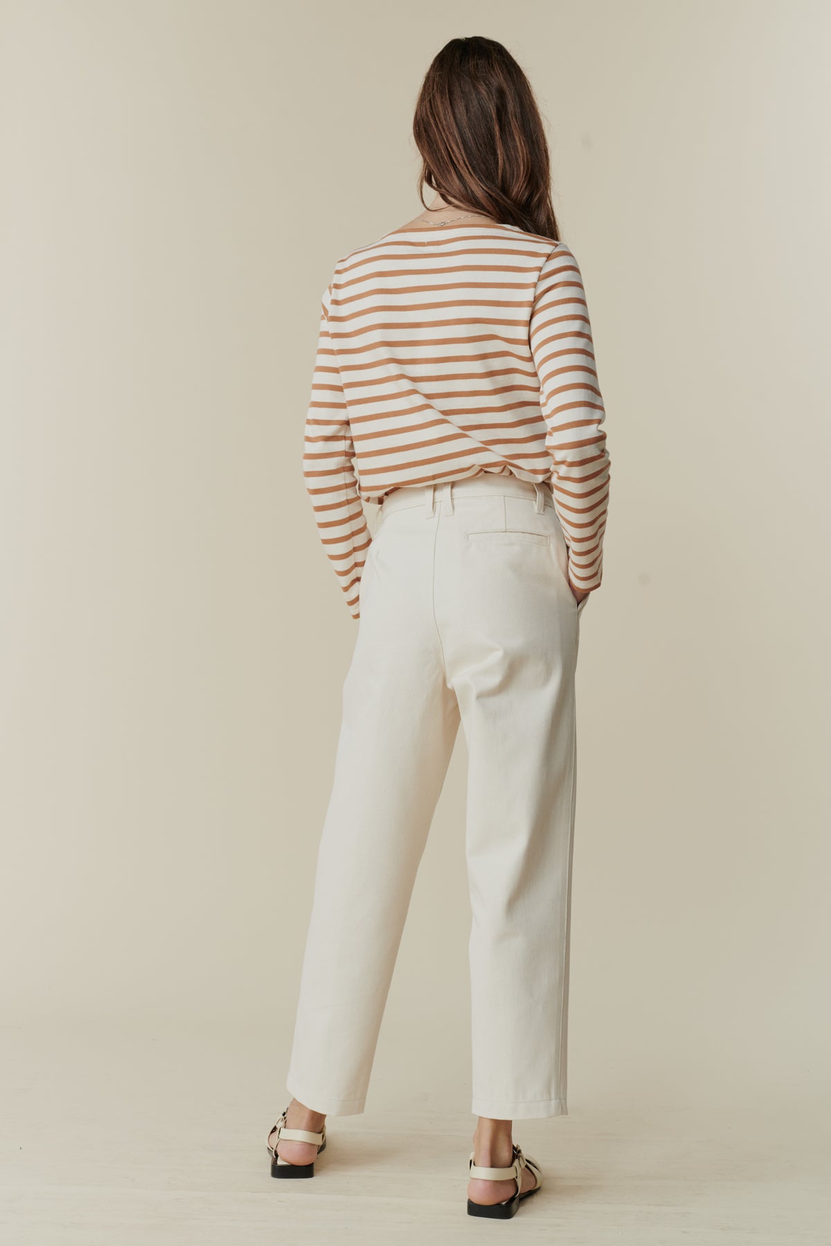 Full body image of back of brunette female wearing women's pleated jeans in ecru