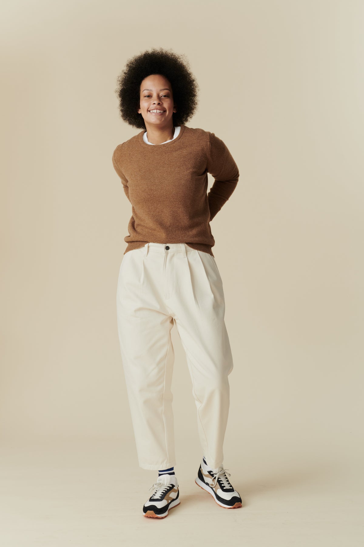 
            Full body shot of black female with afro wearing women&#39;s pleated jean in ecru paired with dark camel lambswool crew neck jumper and Walsh x community clothing trainers