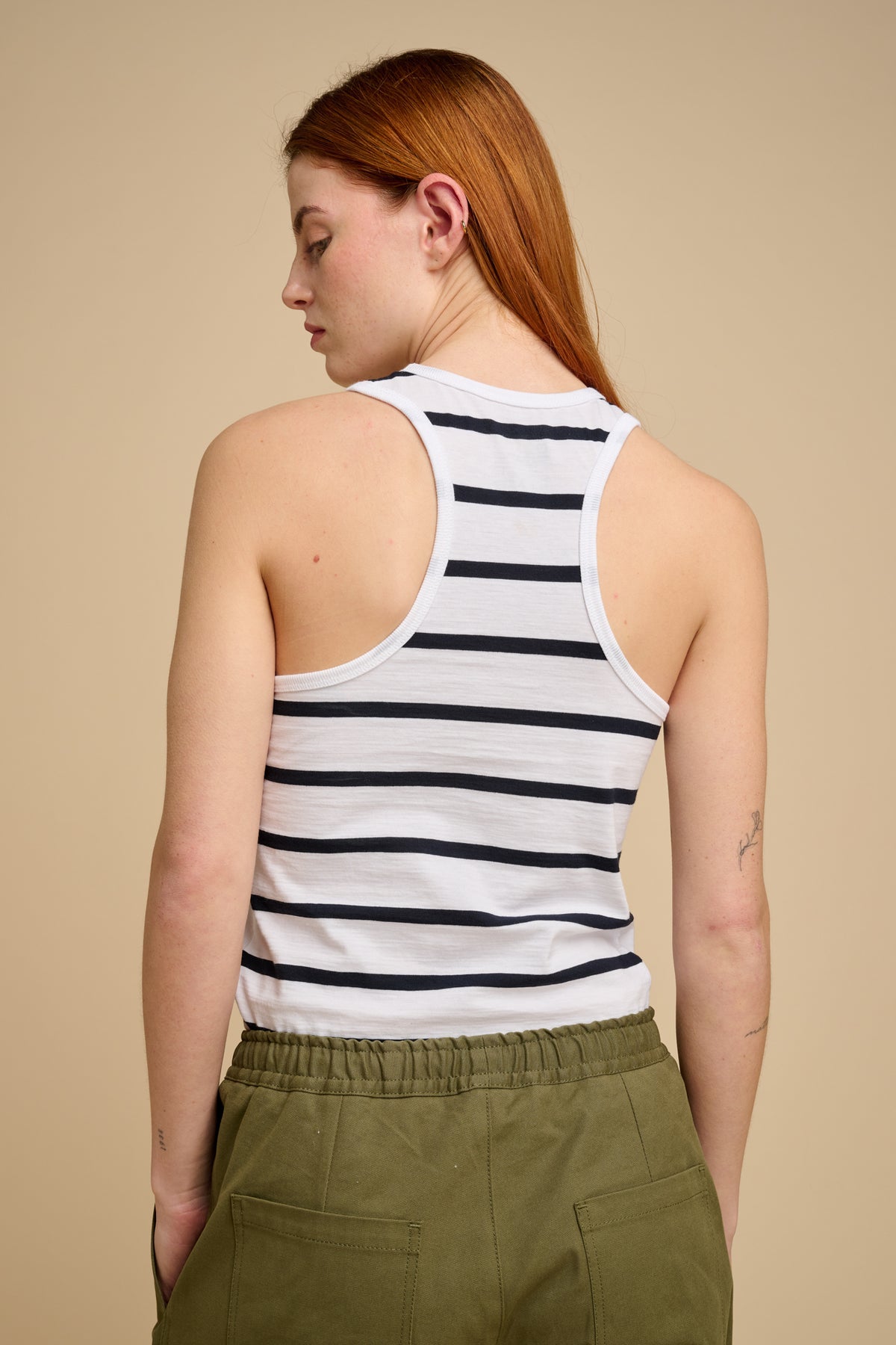 The back of woman wearing a striped racer back vest and green pants against a beige background.