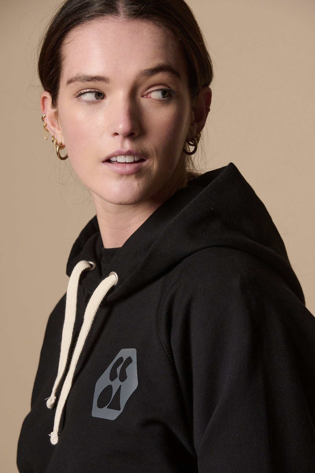 
            Portrait from the side of female wearing women&#39;s raglan hooded sweatshirt plastic free in black with ecru drawstring for the hood. CCOA logo of left of chest