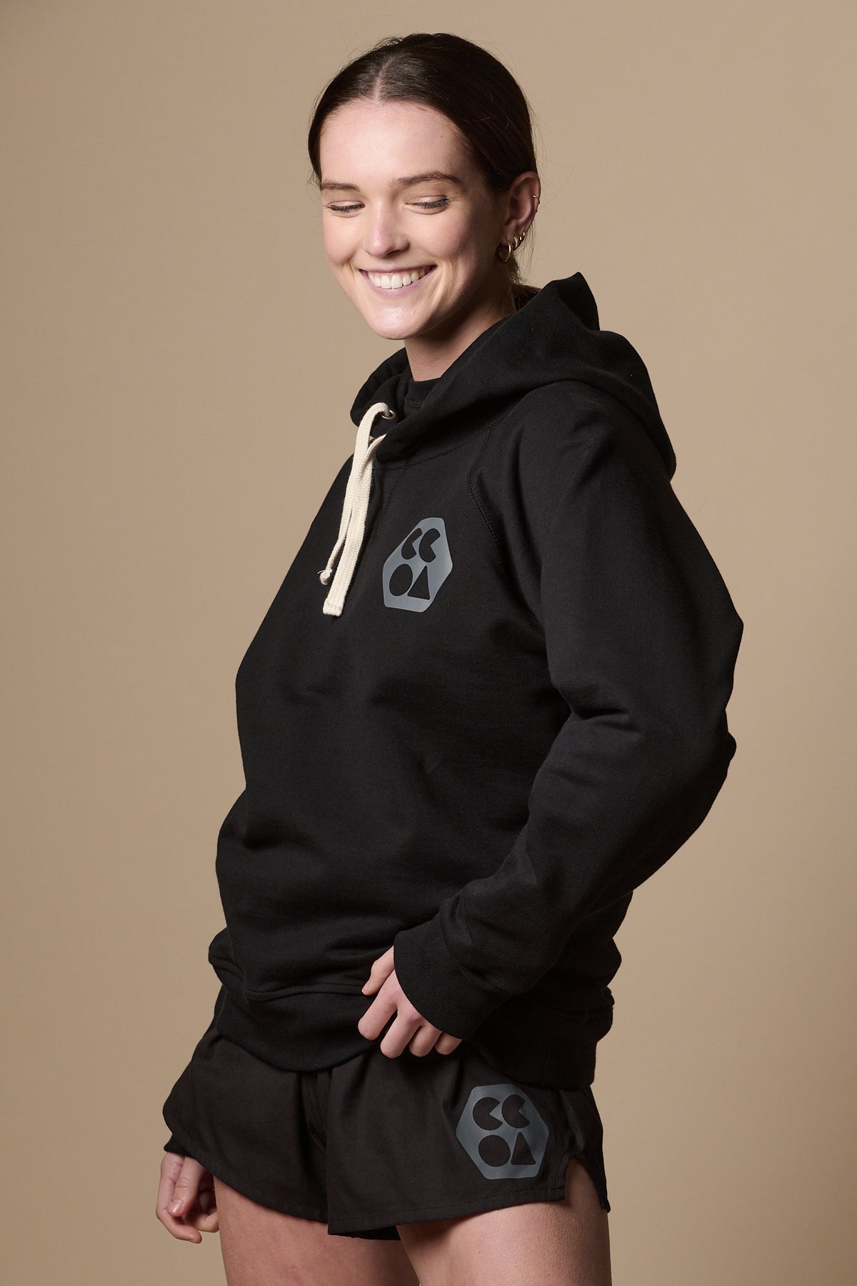 
            Female wearing women&#39;s raglan hooded sweatshirt plastic free in black with ecru drawstring for the hood. CCOA logo of left of chest
