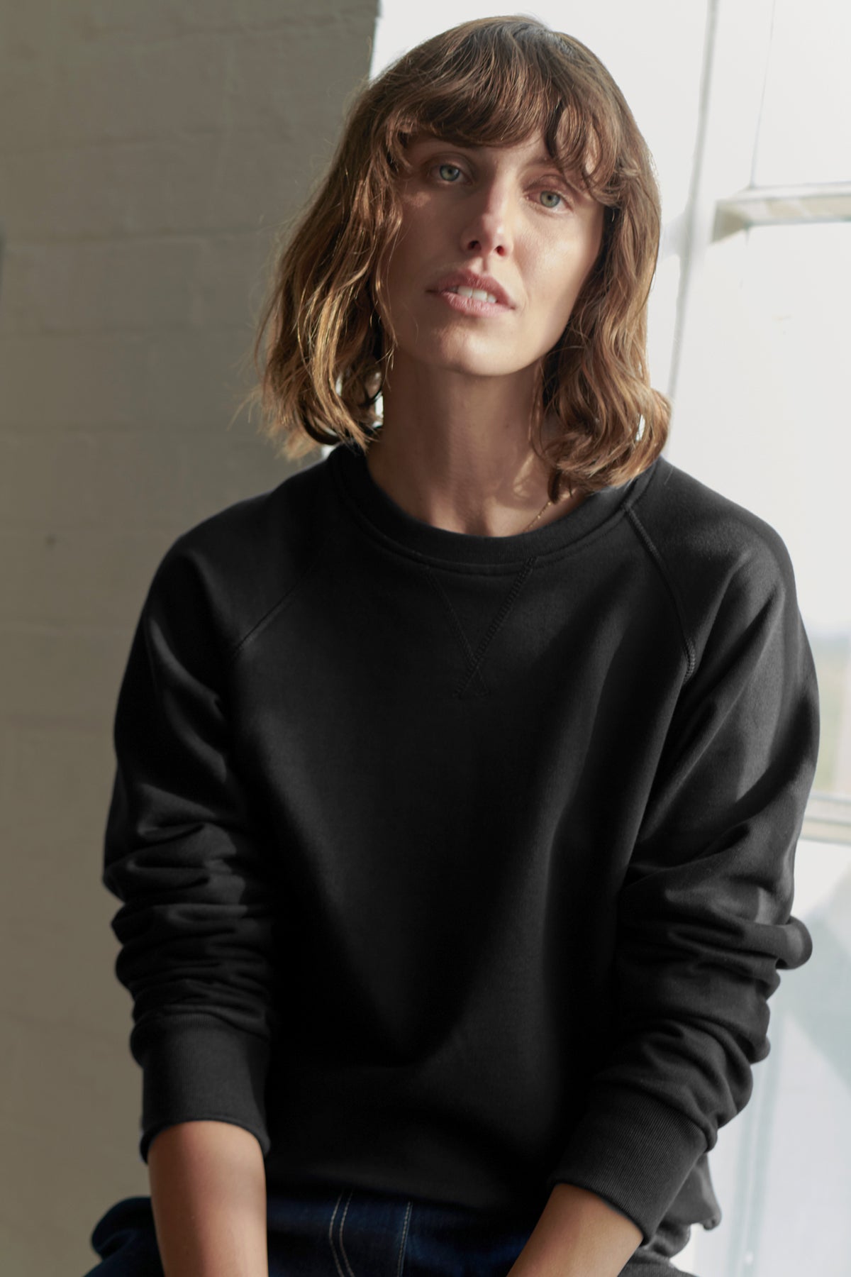 
            Thigh up image of brunette, white female wearing women&#39;s raglan sweatshirt in black with the sleeves pushed up