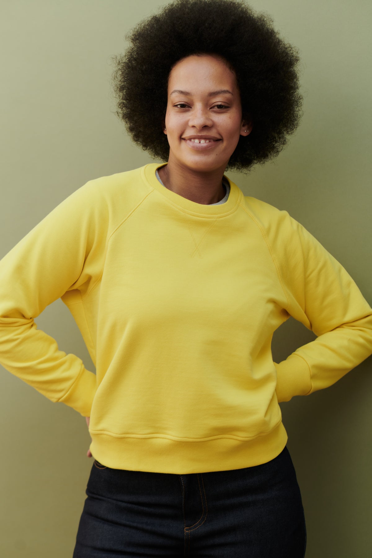 
            Curly haired, smiley female, with hands on hips wearing raglan sweatshirt in canary yellow