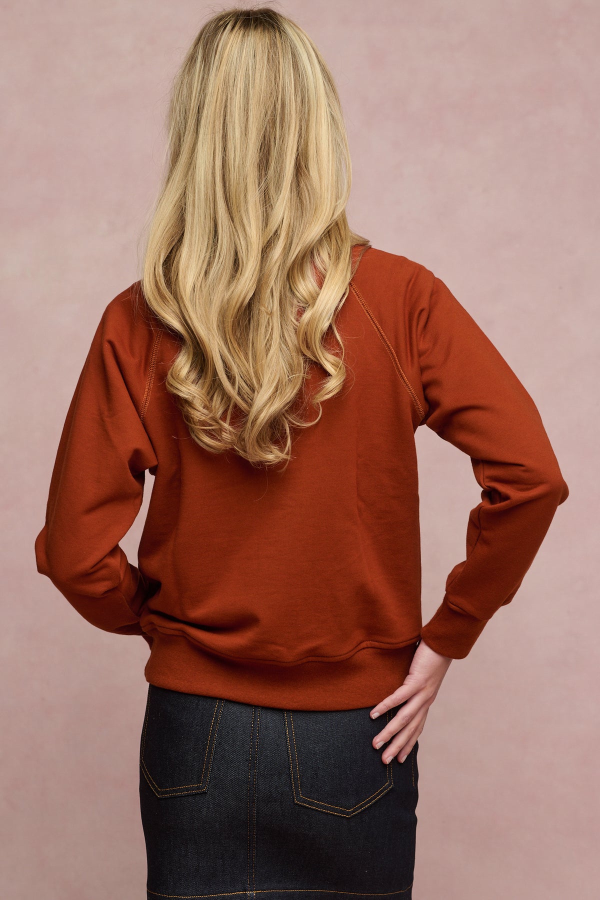 Thigh up image of the back of female with long blonde curled hair wearing raglan sweatshirt in cinnamon