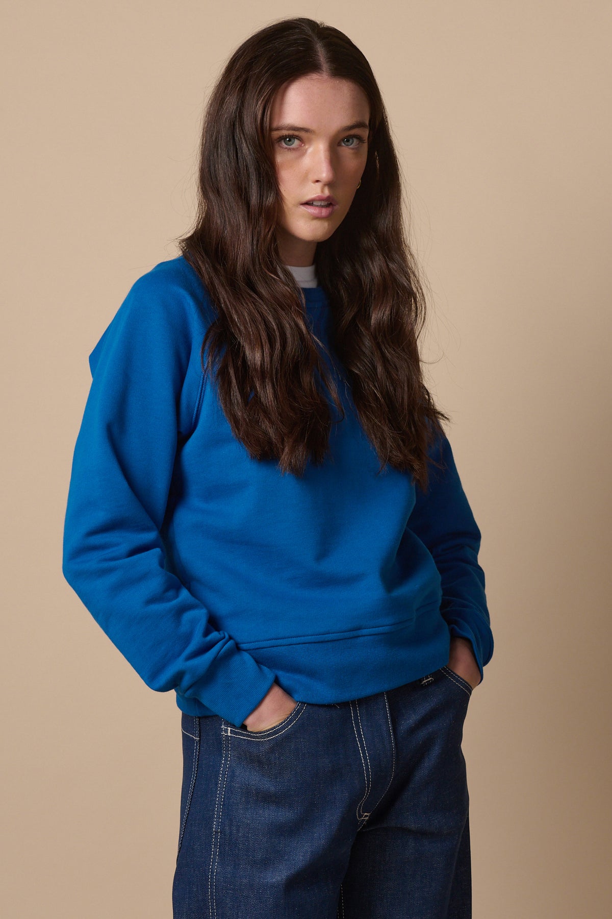 Thigh up image of female wearing raglan sweatshirt in cobalt with hands in jeans pockets