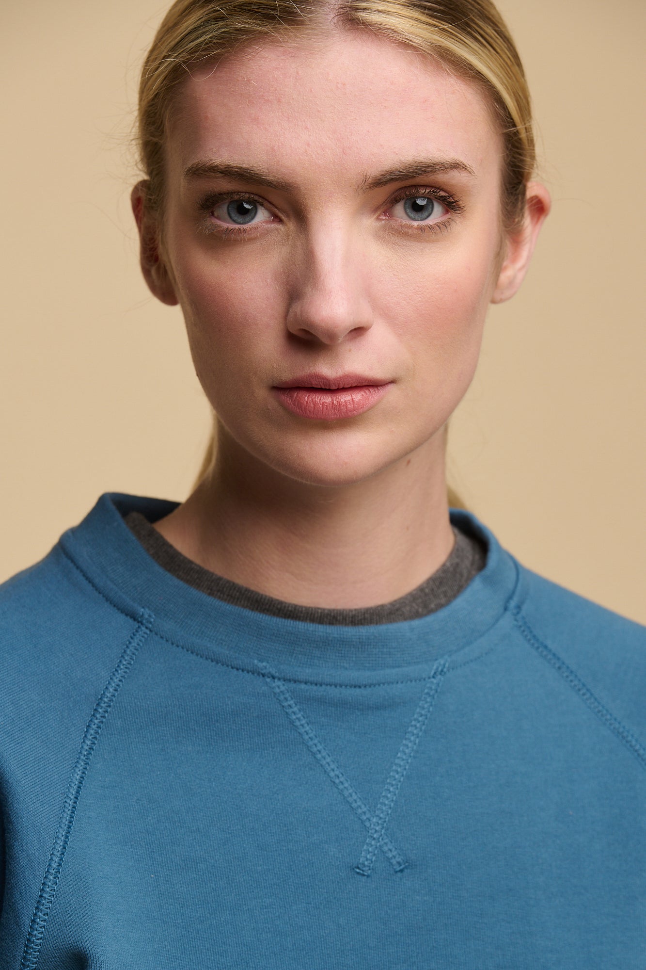 Close up portrait of female with light blue eyes wearing raglan sweatshirt in RAF blue layered over crew neck t shirt in charcoal