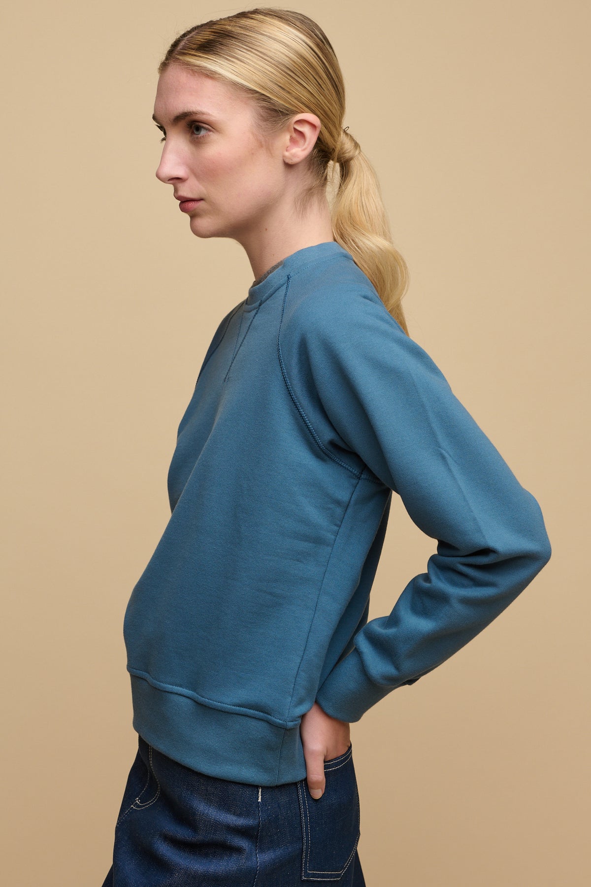 Left side of blonde female wearing raglan sweatshirt in RAF blue with her left hand in back pocket of Bobbie denim midi skirt in blue