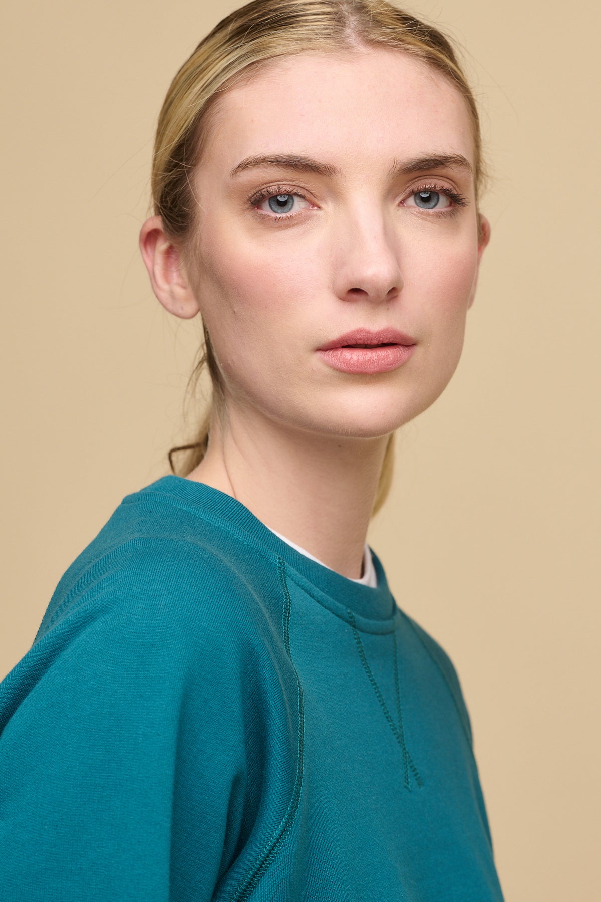Portrait of blonde female with fair skin wearing raglan sweatshirt in teal, layered over crew neck white t shirt