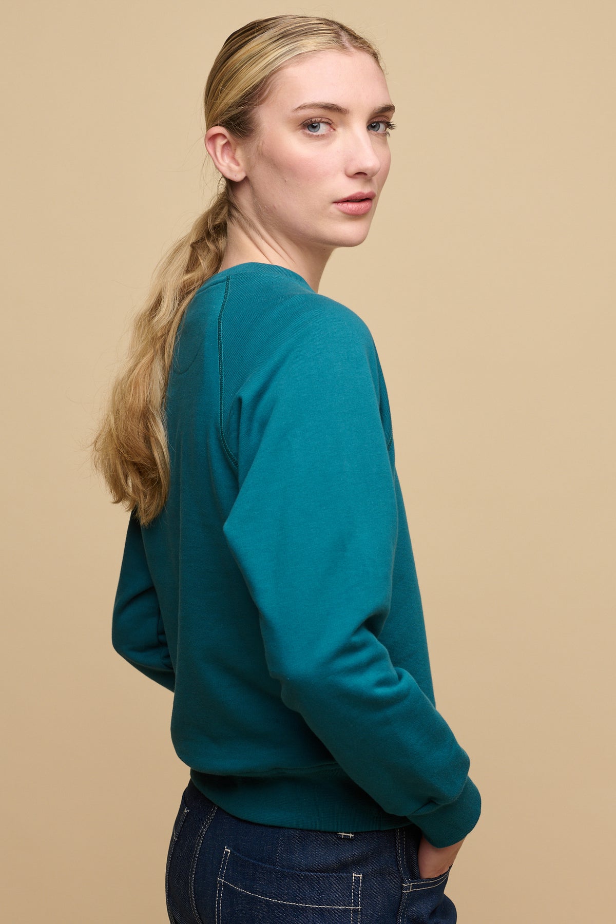 
            The side of female with blonde hair tied in low ponytail wearing raglan sweatshirt in teal paired with work jeans in blue