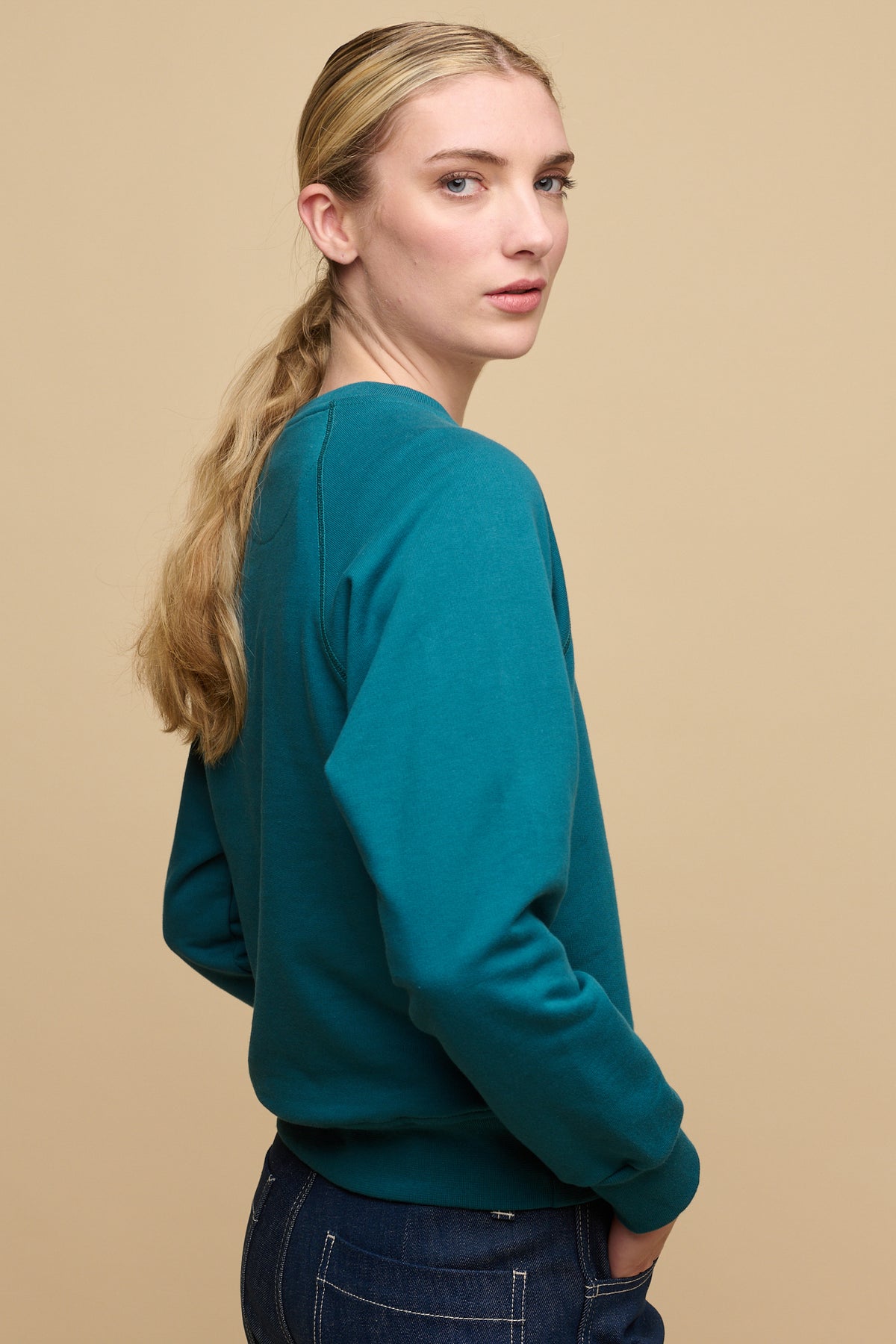 The side of female with blonde hair tied in low ponytail wearing raglan sweatshirt in teal paired with work jeans in blue