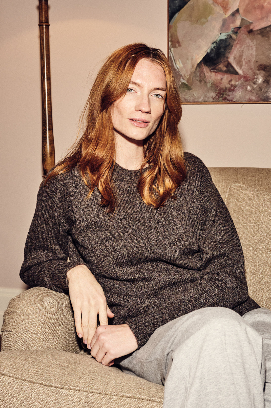 Woman sitting on a couch wearing a brown jumper and light pants, with a neutral background.