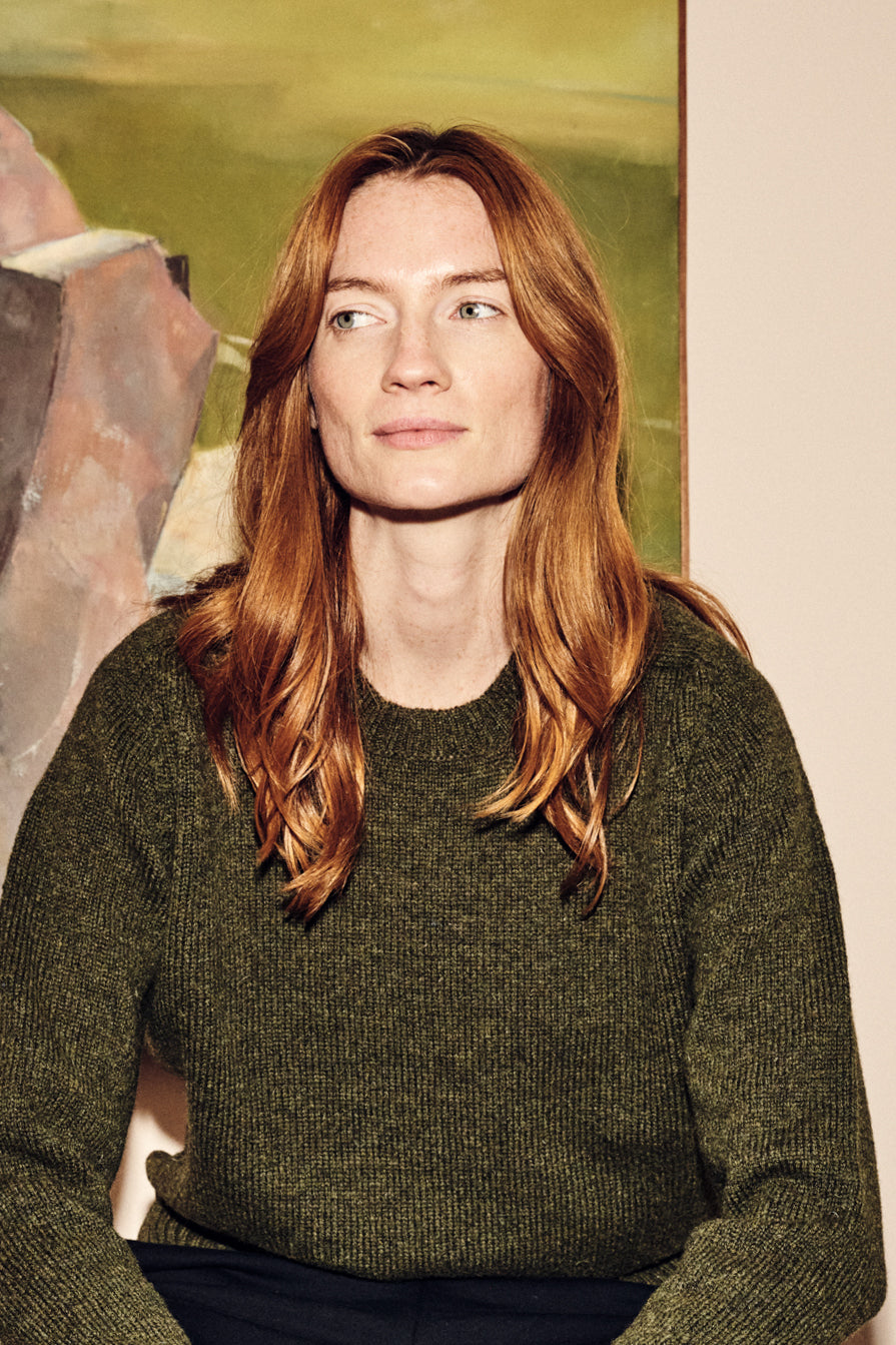 Woman with red hair wearing a green sweater in front of a painting